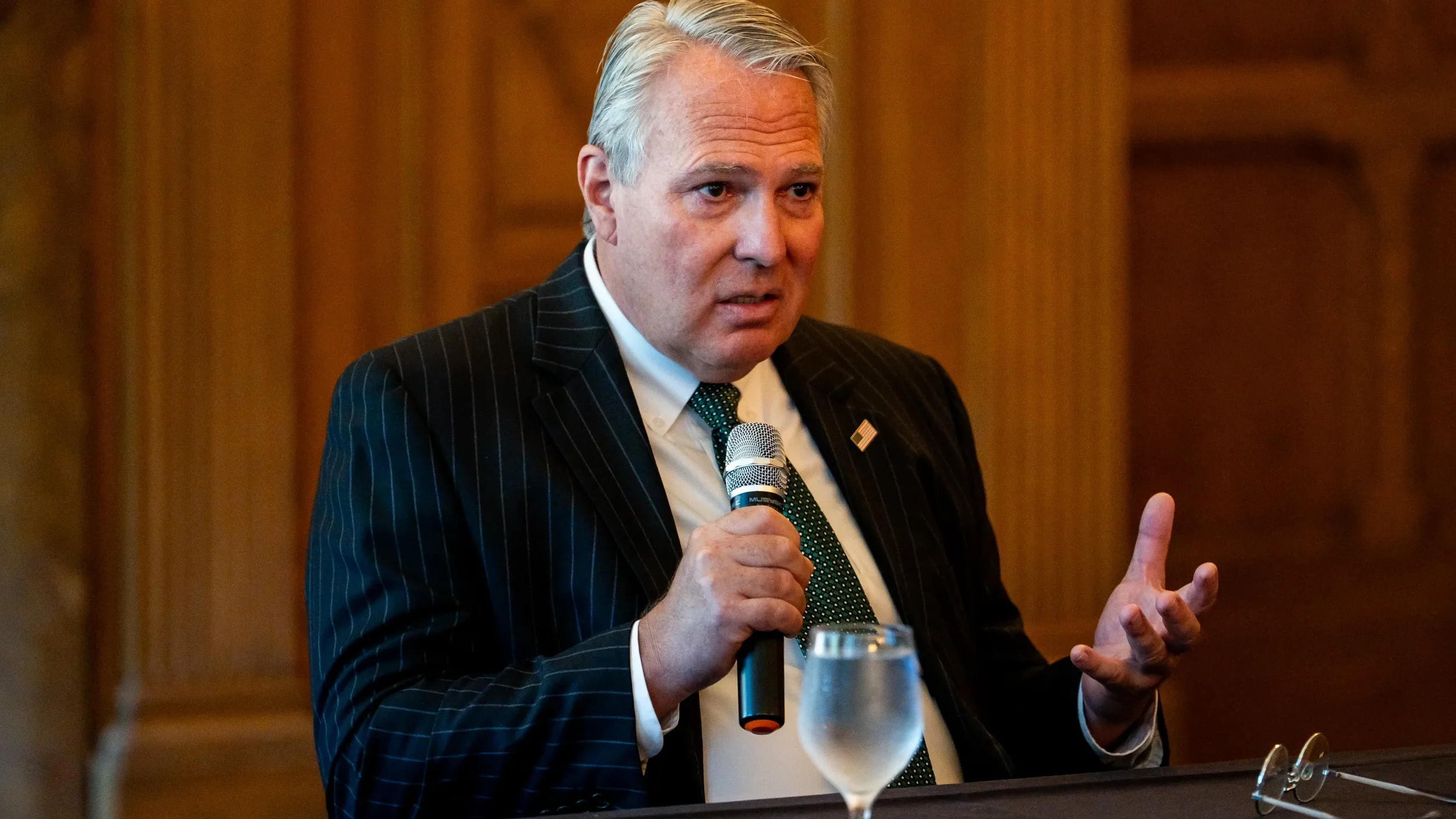 A man with gray hair wearing a dark pinstripe suit, white shirt, and green tie speaking into a handheld microphone at a formal event or conference.