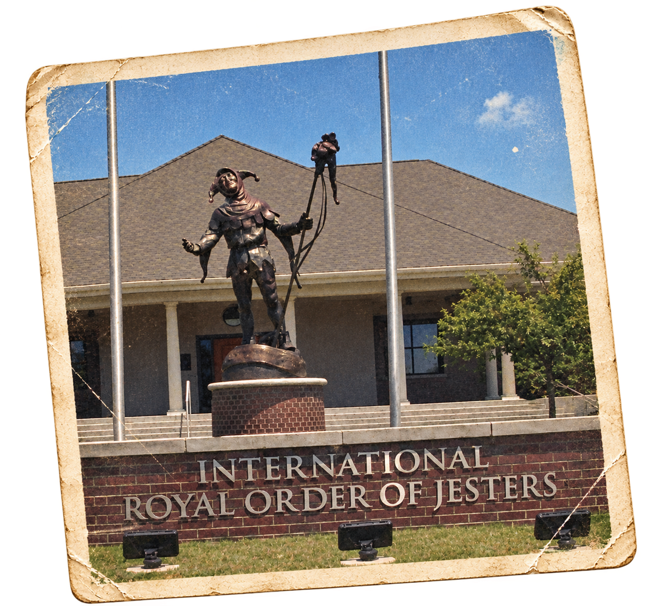 Statue of a jester with juggling clubs on a brick pedestal in front of a building with the sign 'International Royal Order of Jesters' and a house with a porch, trees, and a blue sky.