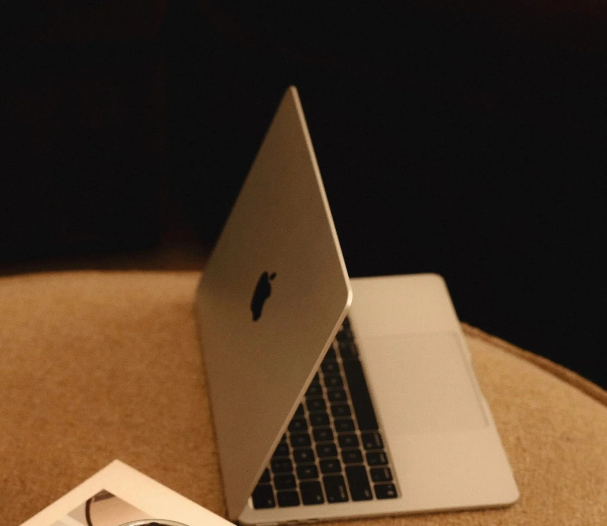 A silver MacBook laptop with a black keyboard is open on a beige table, with some papers and a white object nearby.