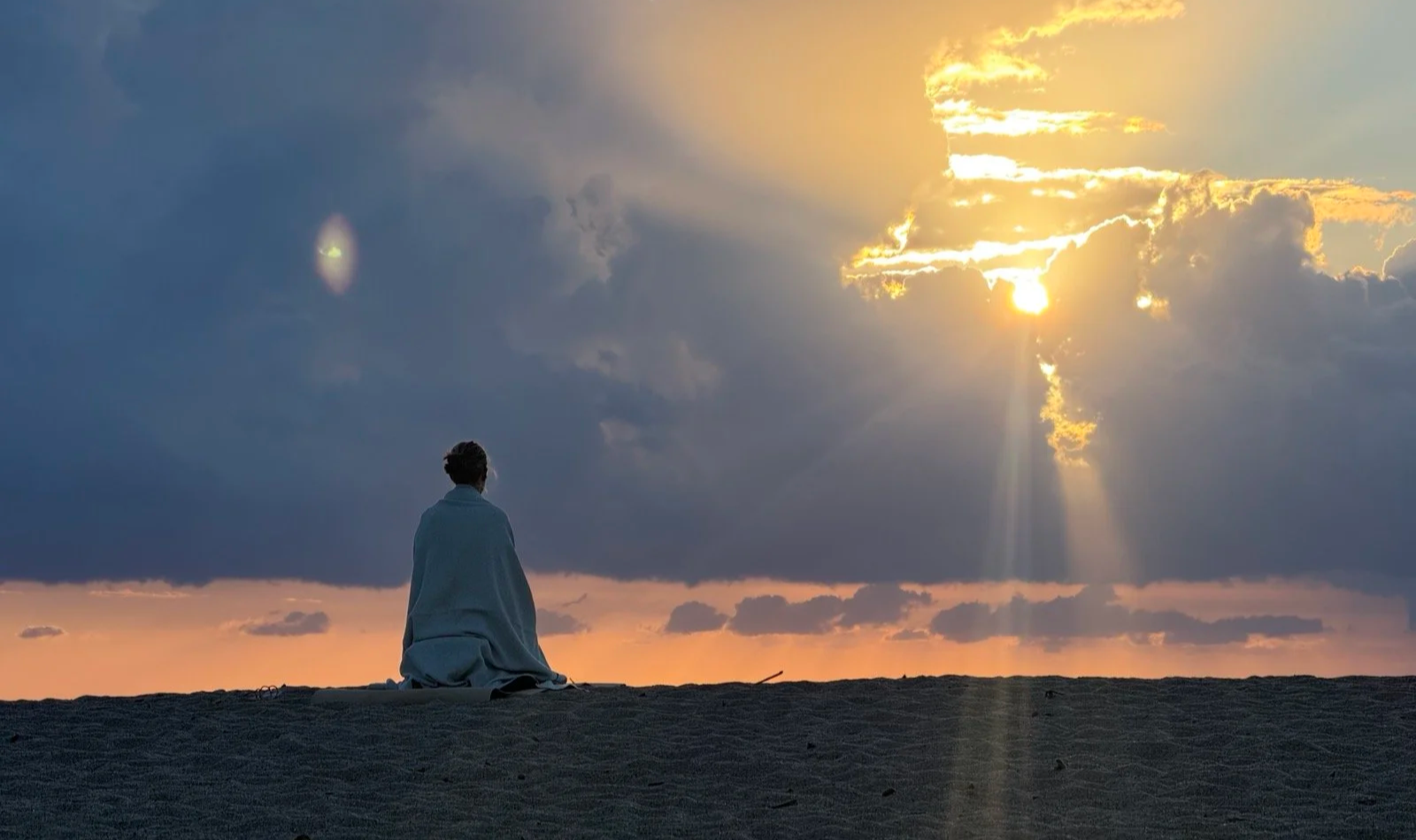 Eine in eine Decke gehüllte Person sitzt bei Sonnenuntergang auf einem Sandstrand und blickt auf den dramatisch bewölkten Himmel.