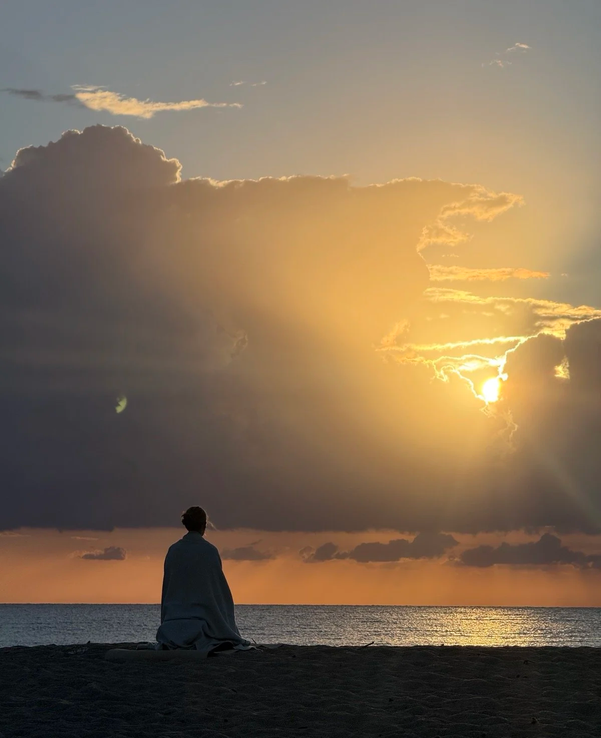 Person in eine Decke gehüllt sitzt am Strand und betrachtet den Sonnenuntergang über dem Meer.