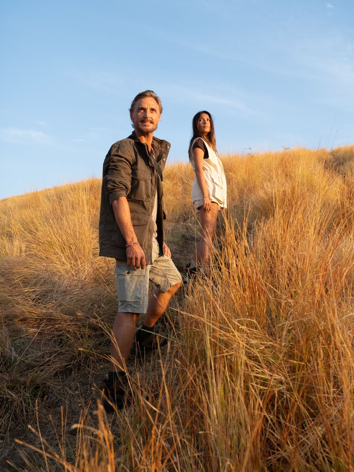 Man and woman standing in a sunlit field of tall golden grass under a clear blue sky