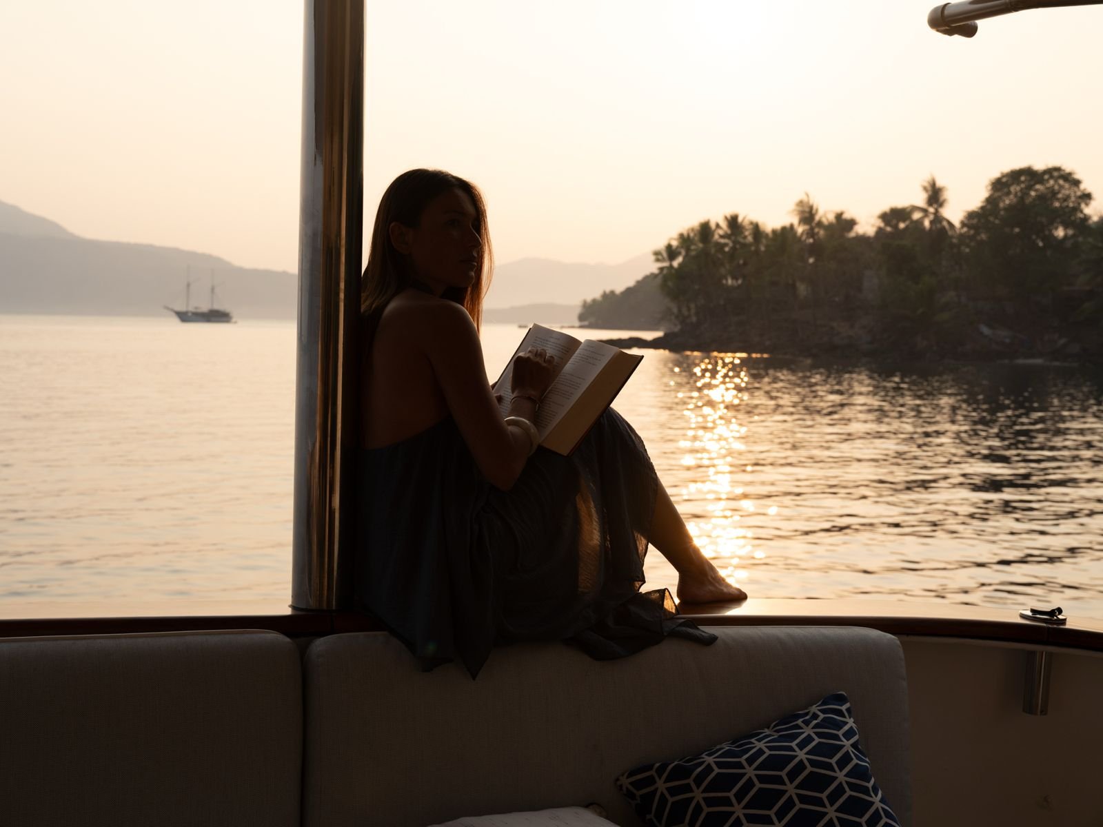 Woman reading a book on a yacht at sunset overlooking tropical islands and calm water