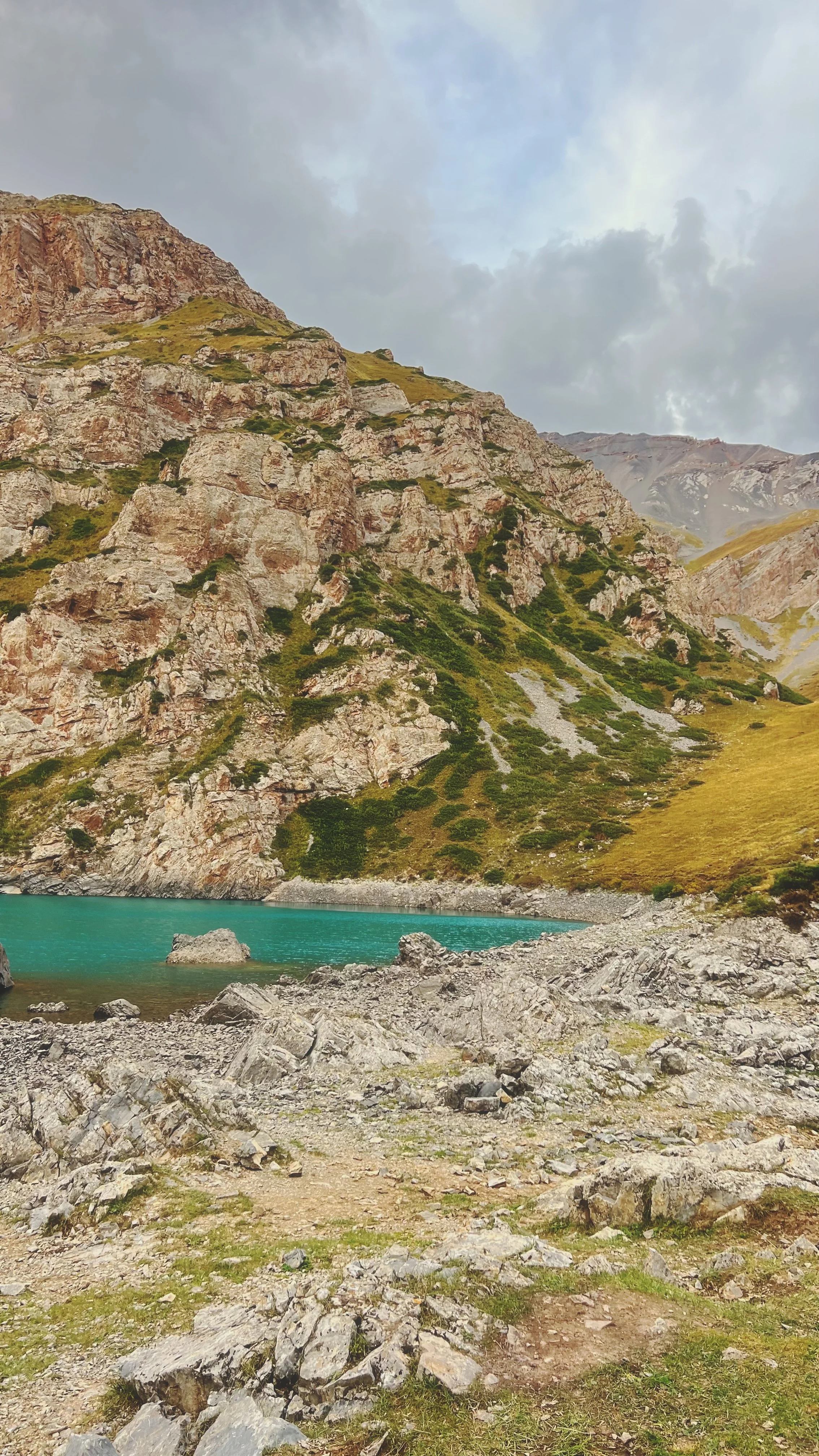 Photo of a mountain with rocky cliffs and green vegetation near a body of water with a rocky shoreline, under a cloudy sky.