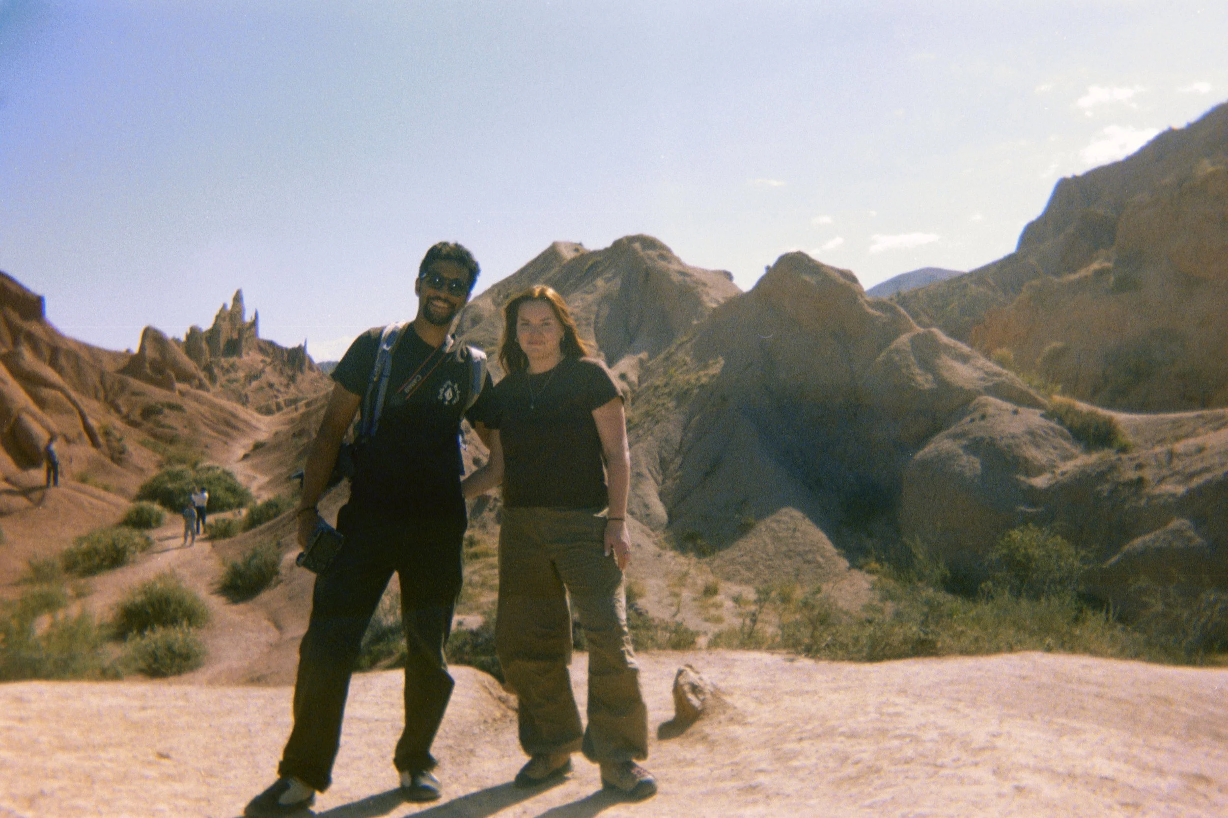Two people standing in a rocky desert landscape with hills and formations in the background, walking trail, clear blue sky, sunny weather.
