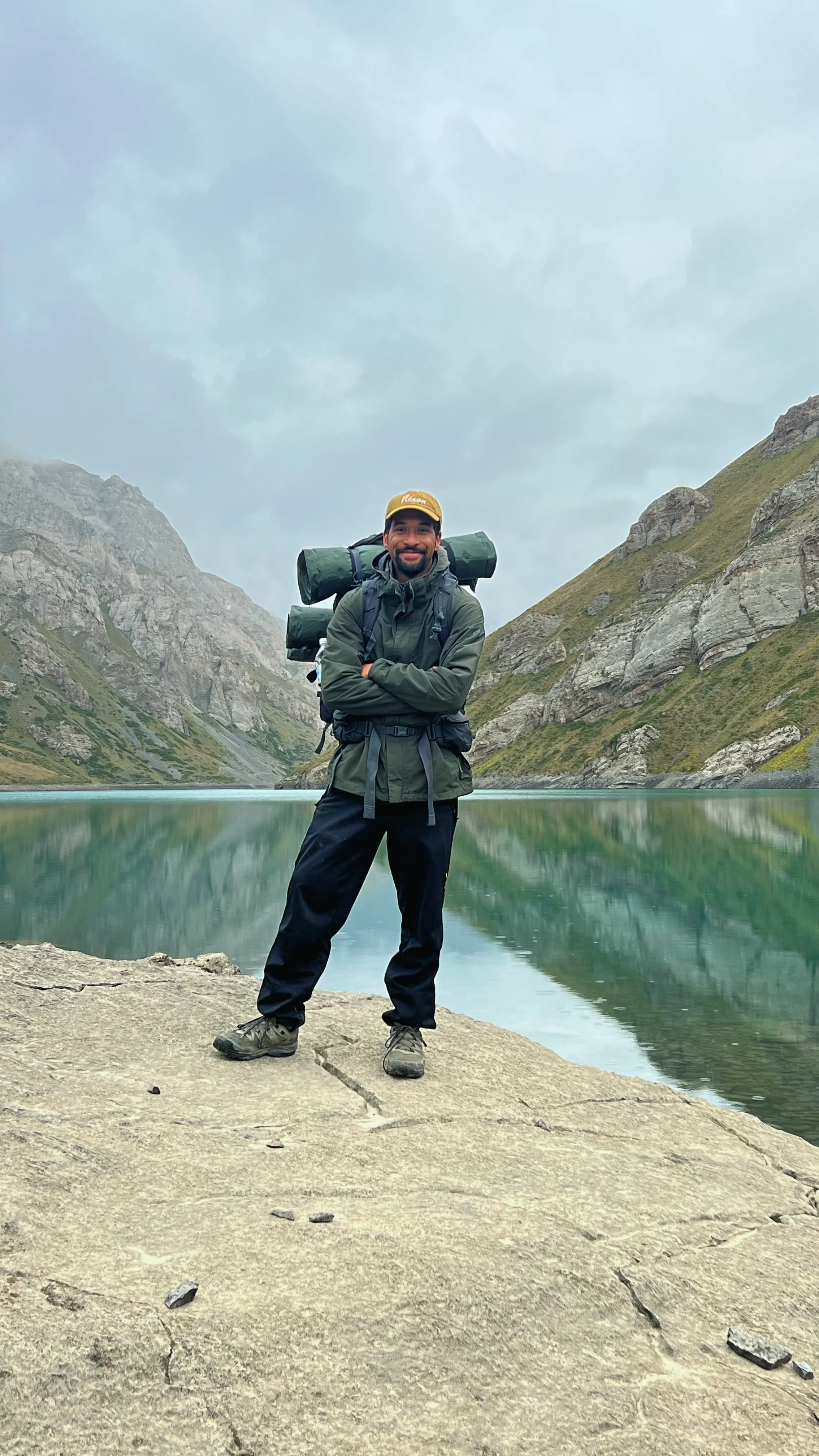 A man standing with arms crossed in front of a mountain lake, wearing hiking gear, with a backpack and a yellow cap, surrounded by steep rocky mountains and cloudy sky.
