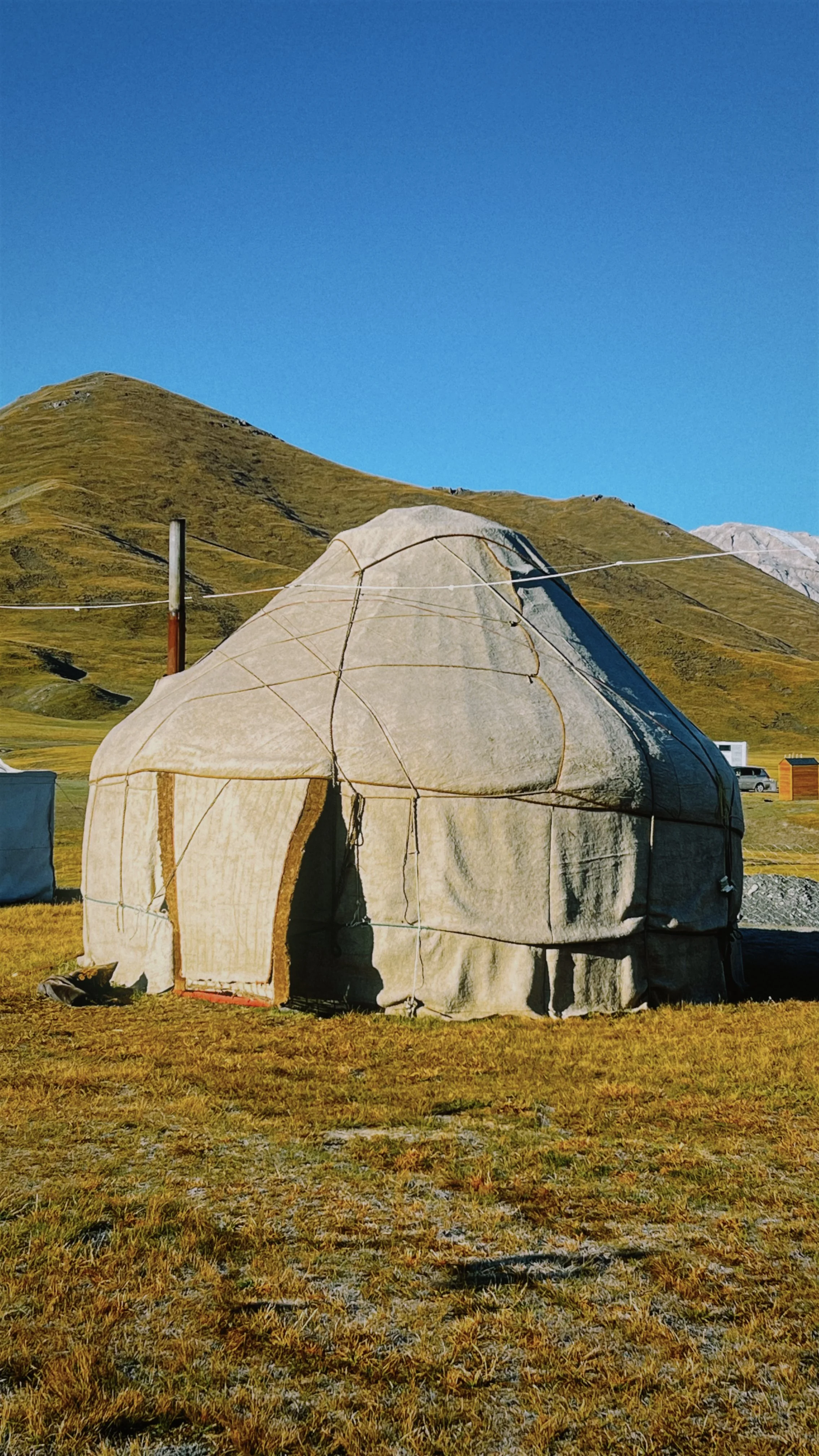 A large beige yurt pitched on grassy ground with mountains in the background and a bright blue sky overhead.
