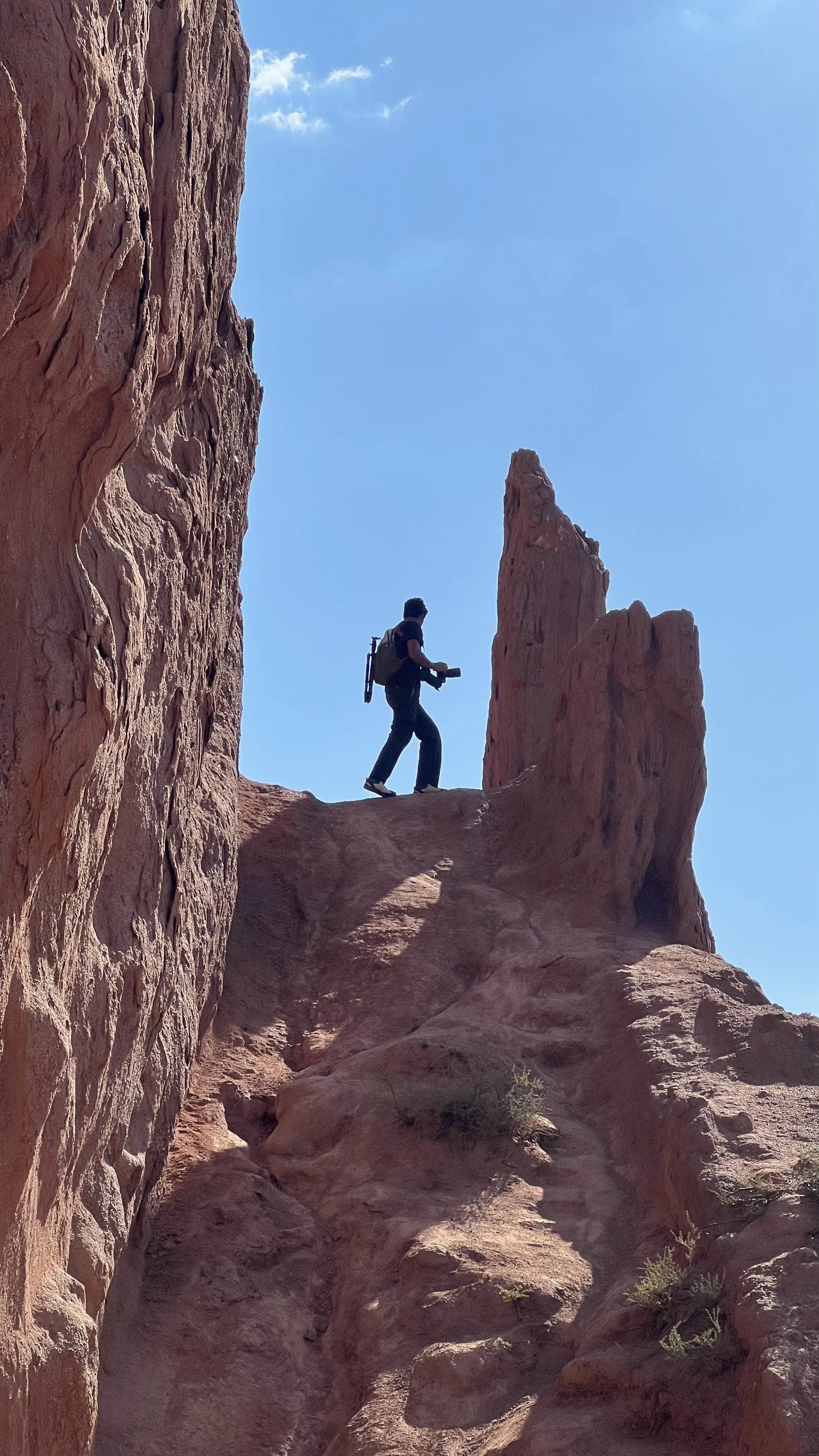 A person with a backpack walking on a rocky, desert landscape with tall red rock formations under a clear blue sky.