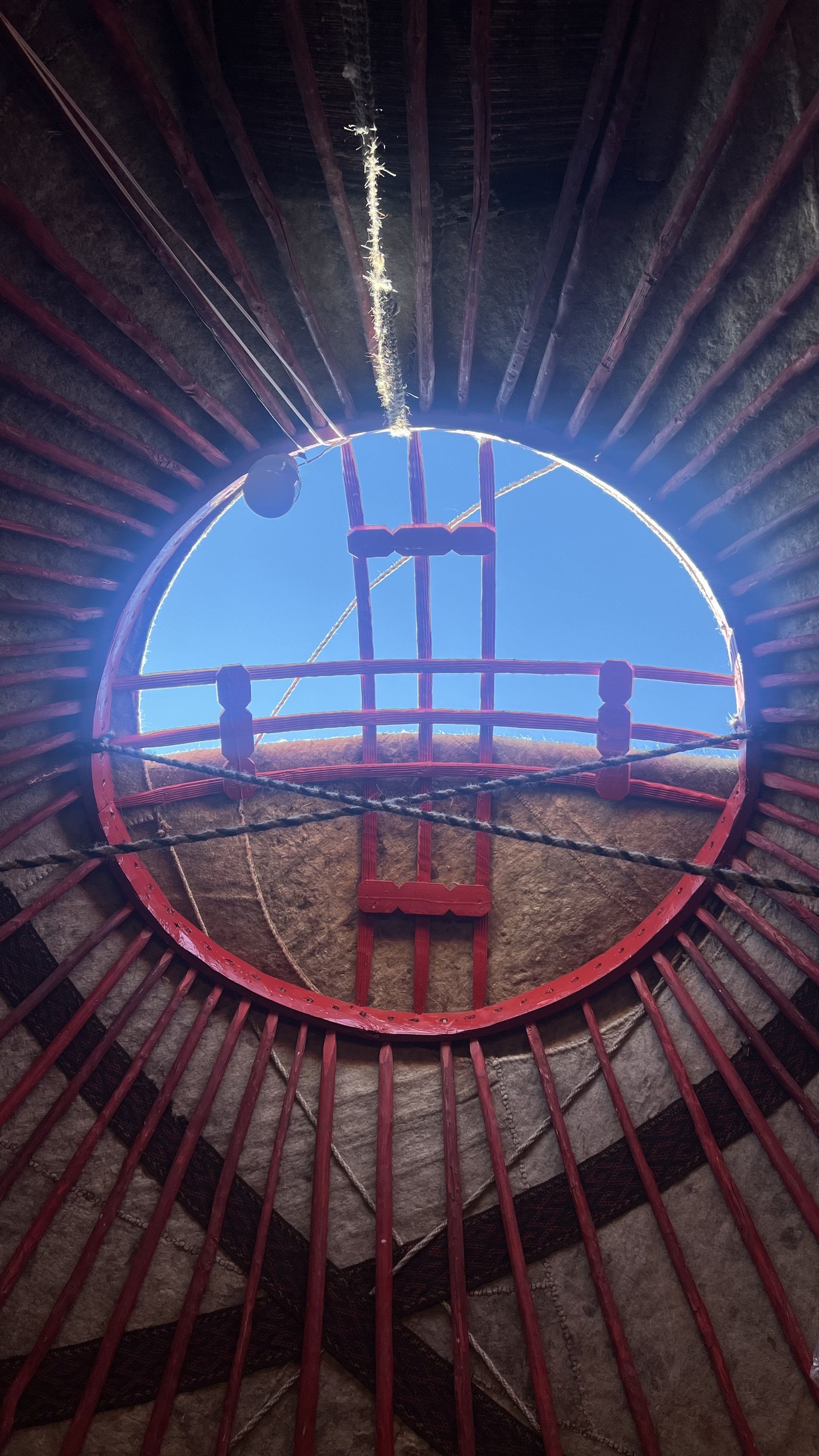 View looking up inside a large bell tower, showing red wooden supports and ropes, with a circular window at the top through which blue sky and sunlight are visible.