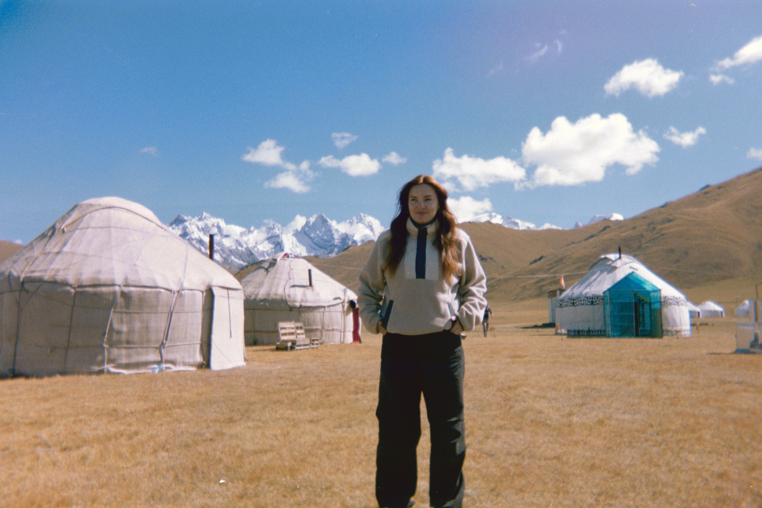 A woman with long hair standing outdoors in front of yurts and snow-capped mountains under a blue sky with clouds.
