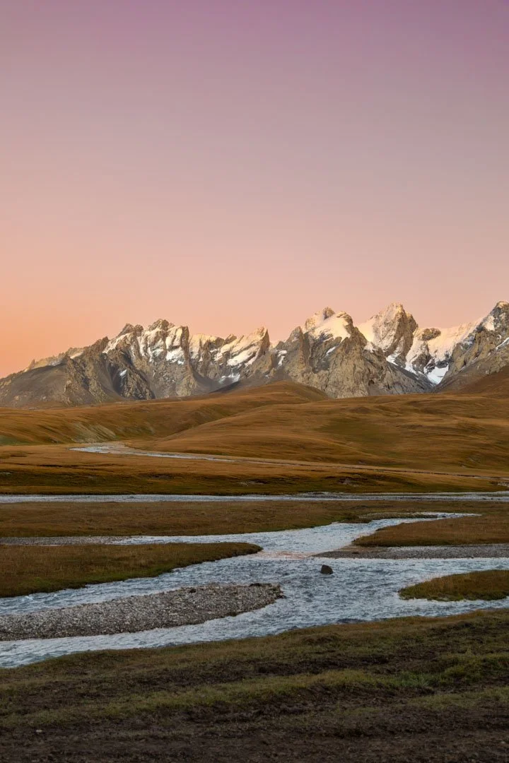 Landscape of snow-capped mountains at sunset, with rolling grassy hills and a winding stream in the foreground.