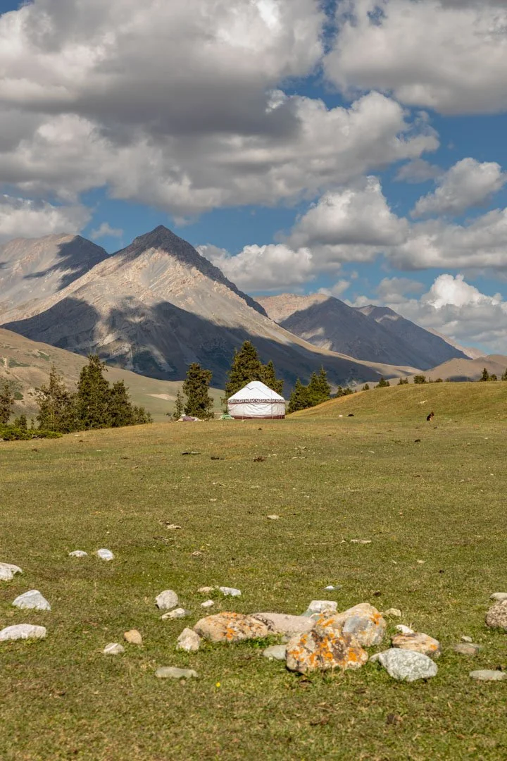 Scenic landscape with a grassy field, scattered rocks, a white tent, trees, mountains in the background, and a partly cloudy sky.