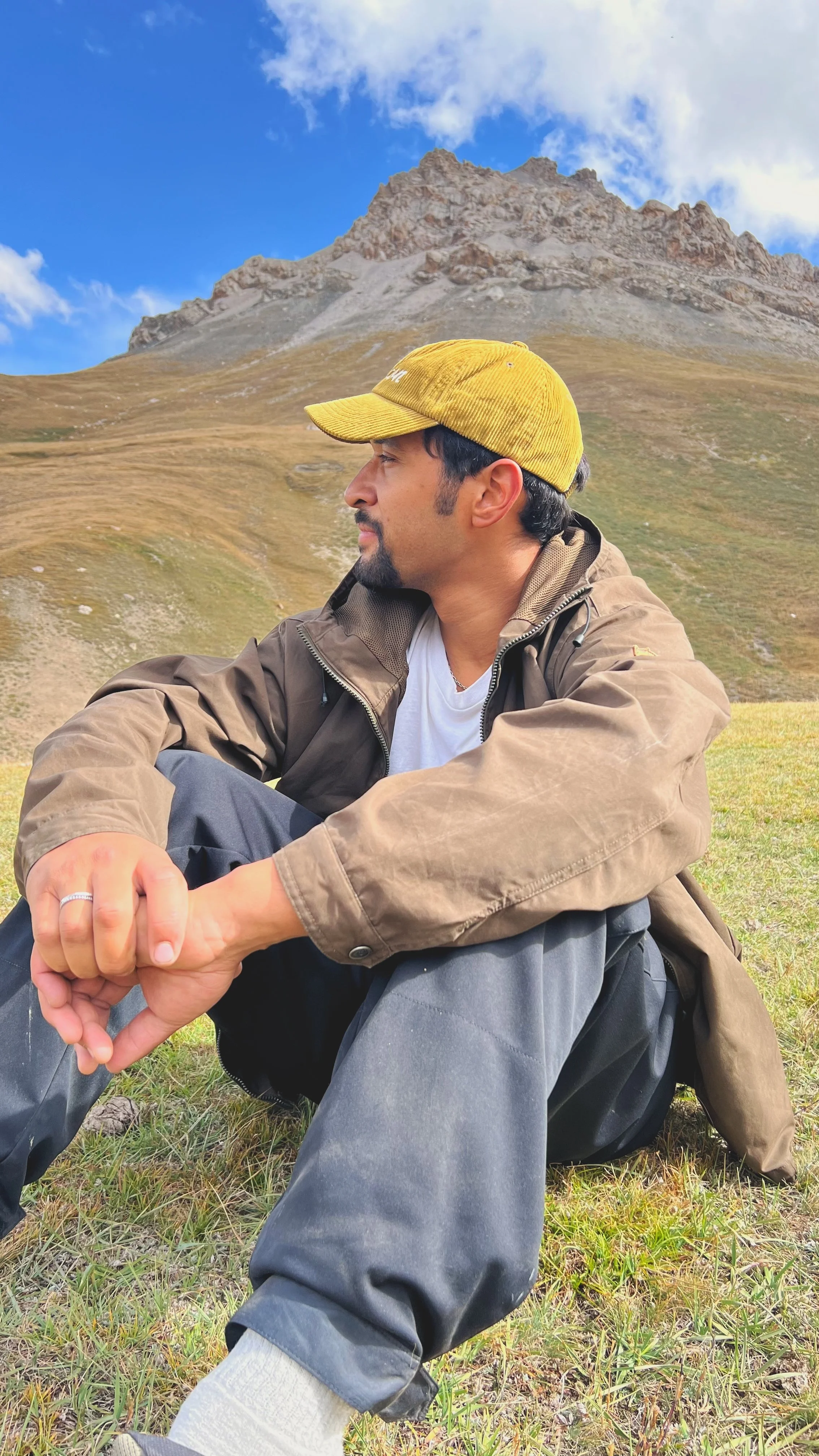 A man sitting on grass in a mountain landscape, wearing a yellow cap, brown jacket, white t-shirt, and gray pants, with mountains and a partly cloudy sky in the background.