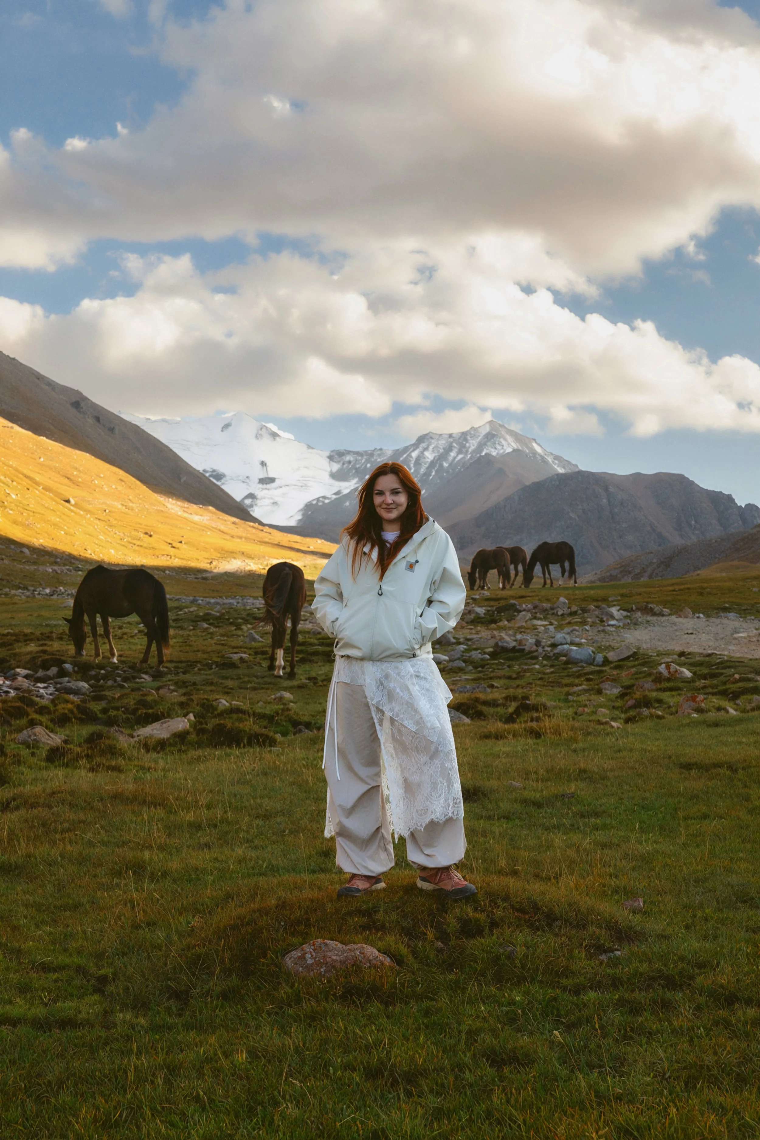 A young woman with red hair wearing a white jacket and beige wide-leg pants standing on a grassy field with mountains and snow-covered peaks in the background, and cows grazing nearby under a partly cloudy sky.