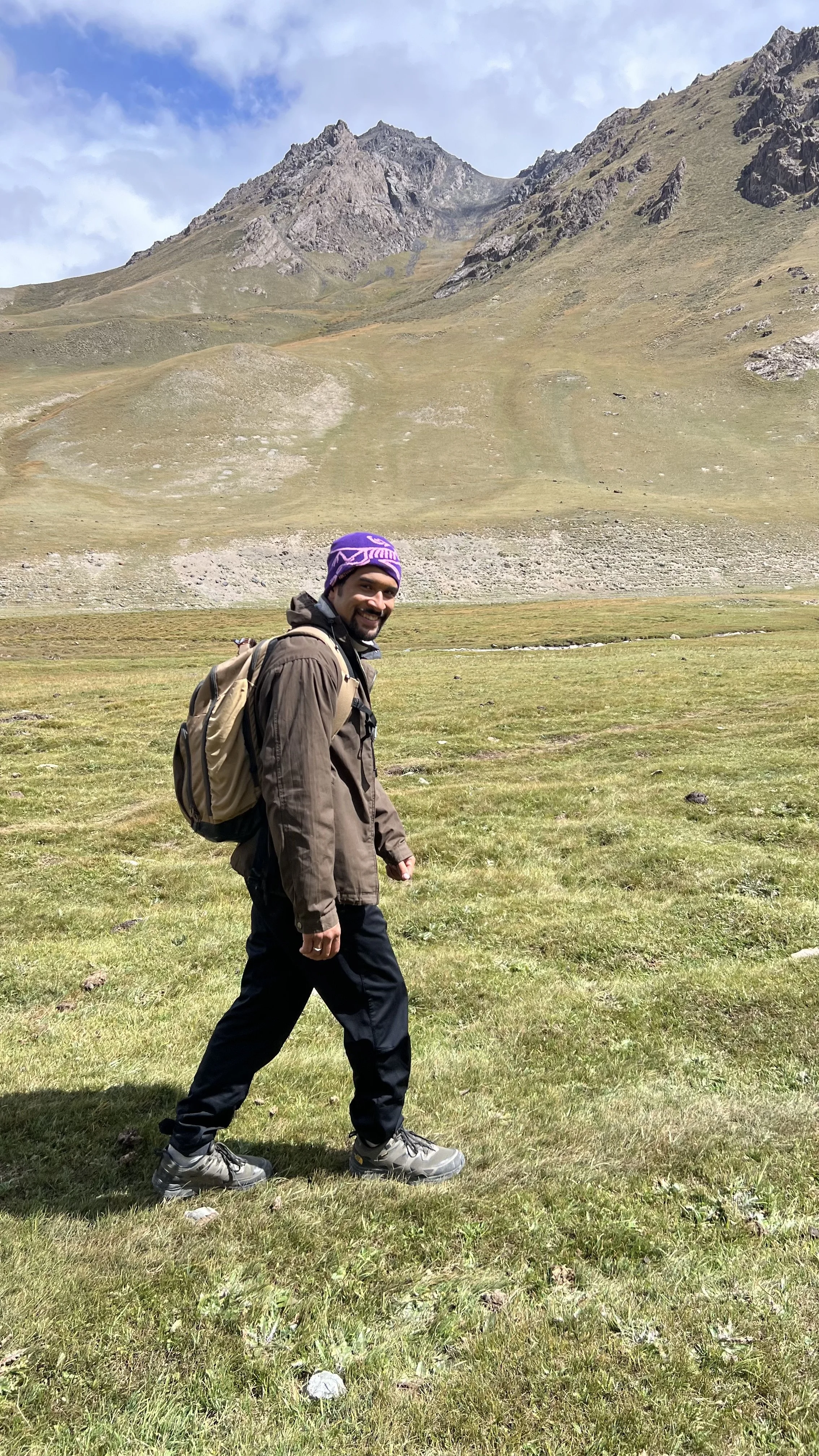 A man with a backpack hiking in a grassy field with mountains and a partly cloudy sky in the background.