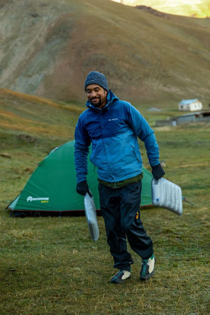A man in outdoor gear carrying foam sleeping pads on a grassy campsite with a green tent and rolling hills in the background.