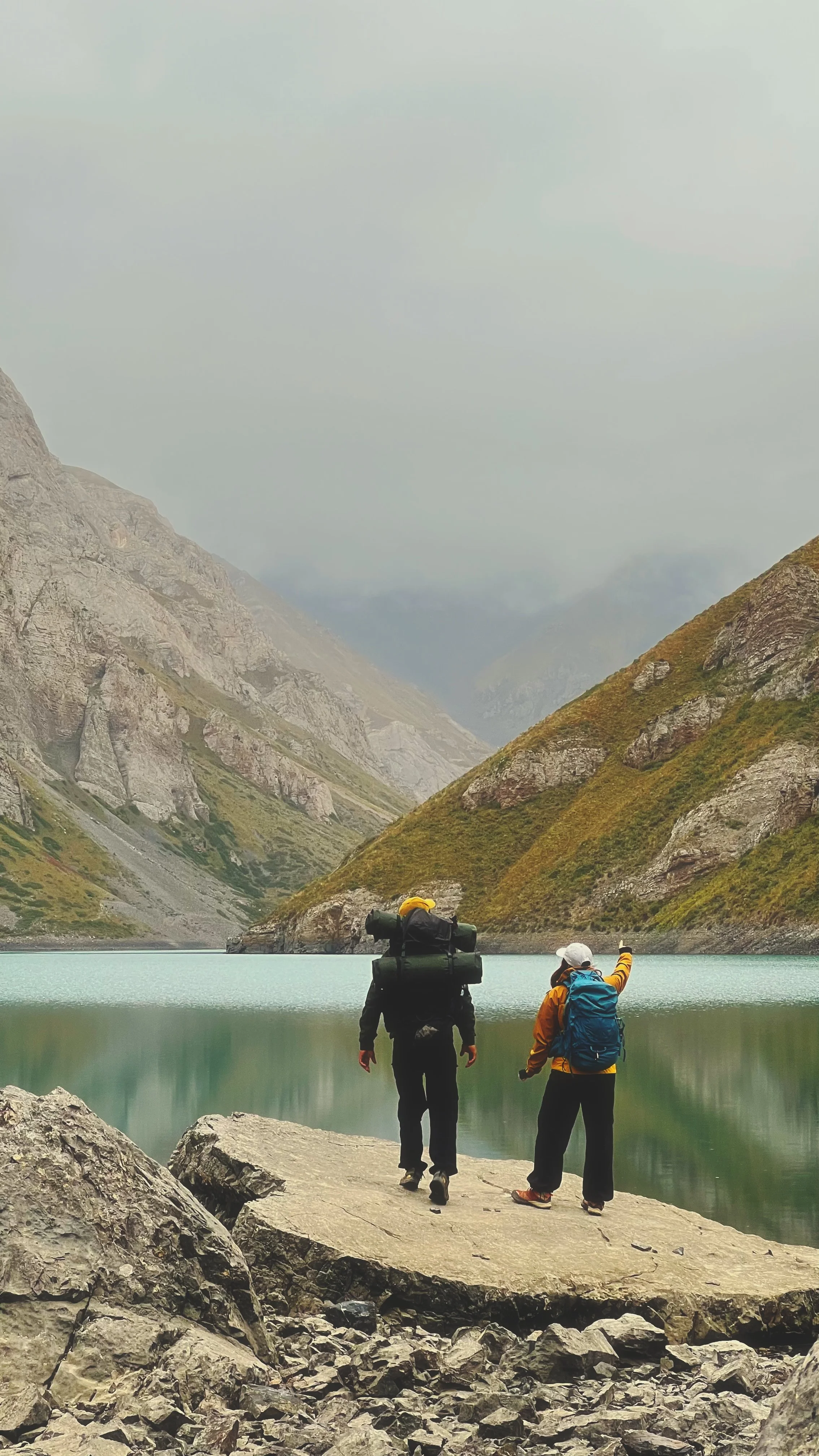 Two hikers with backpacks and gear standing on rocks by a mountain lake surrounded by steep mountain slopes.