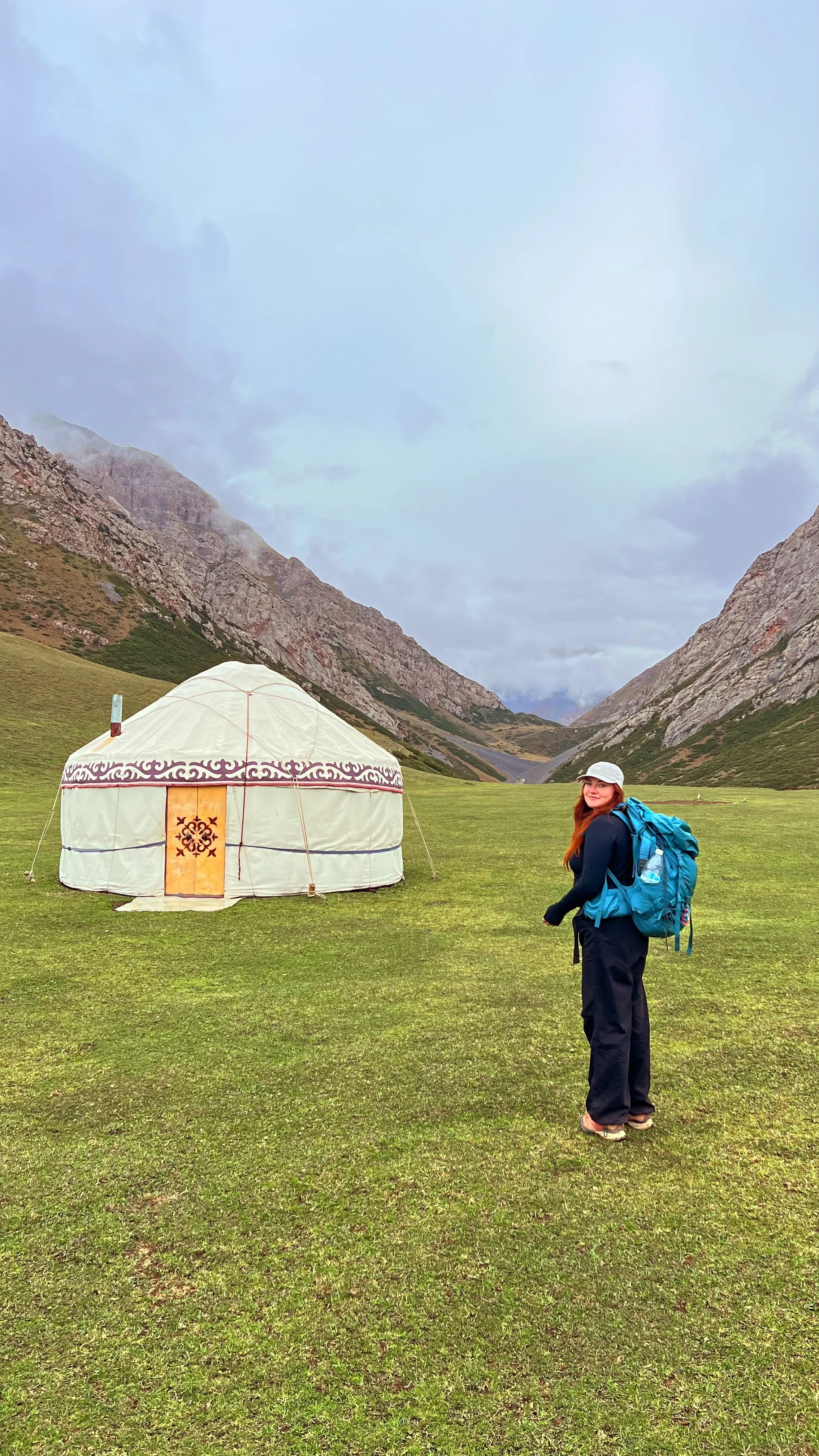 A woman hiking with a large blue backpack in a lush green field, near a traditional yurt, with mountain peaks and a cloudy sky in the background.