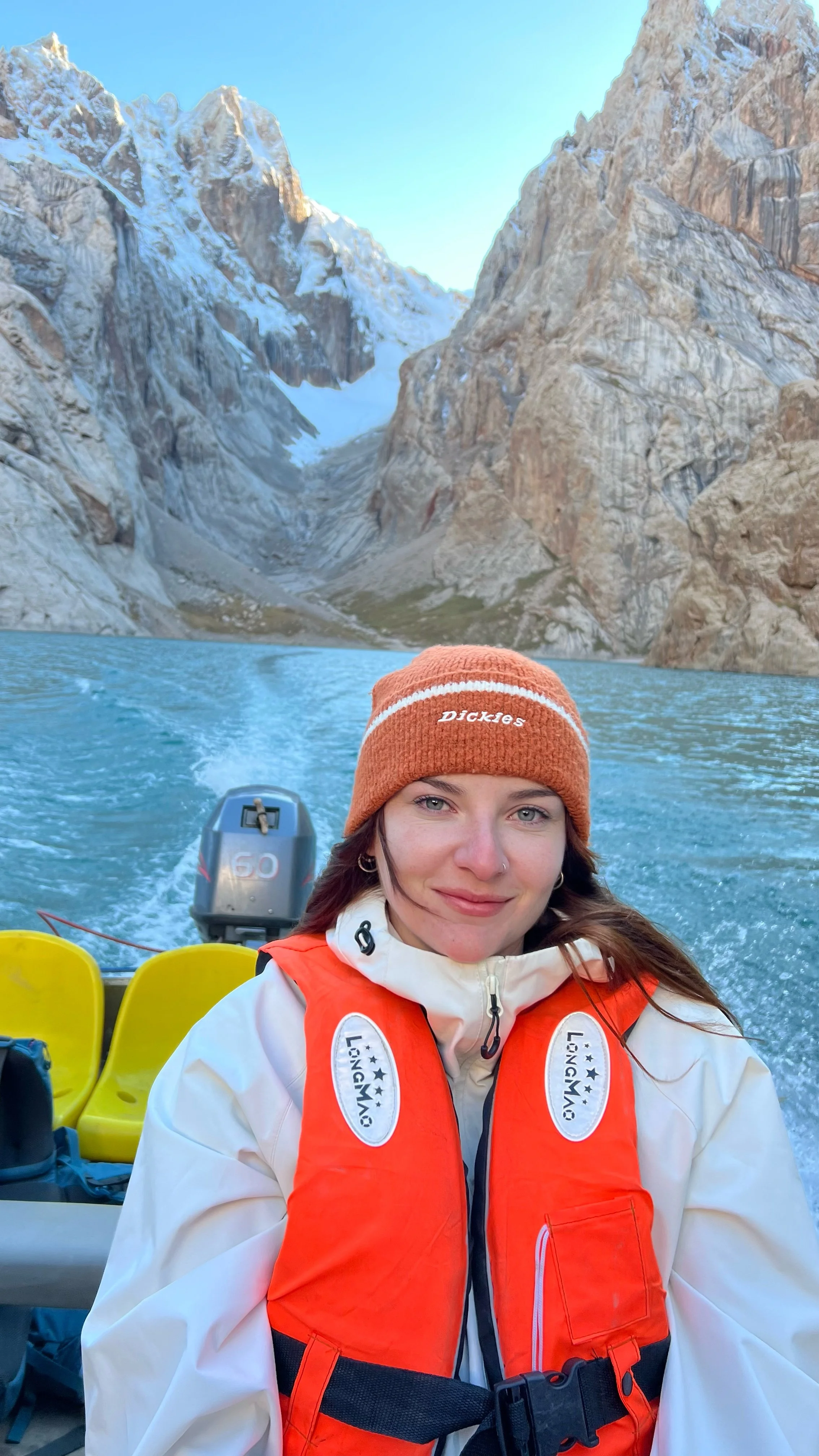 A woman with light skin, blue eyes, brown hair, wearing a tan beanie with 'Dickies' logo and an orange life jacket on a boat, smiling at the camera, with a mountain lake and snowy peaks in the background.