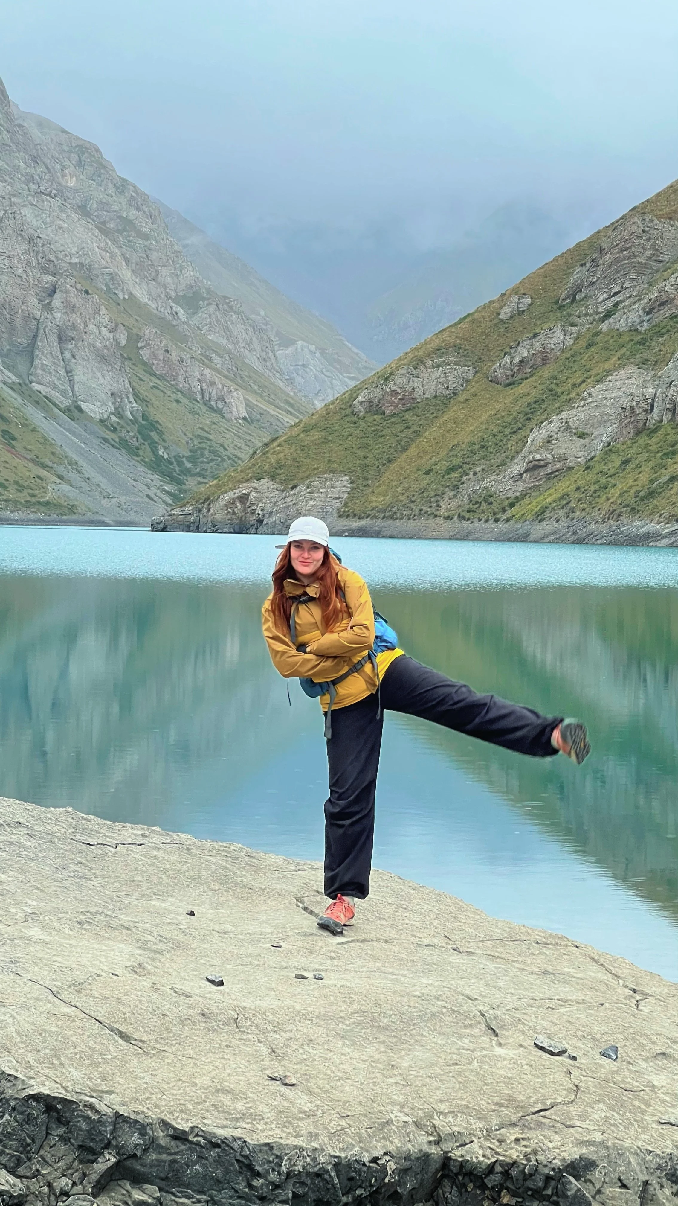 Woman in outdoor clothing posing playfully by a calm mountain lake with tall, rocky, green mountains in the background.