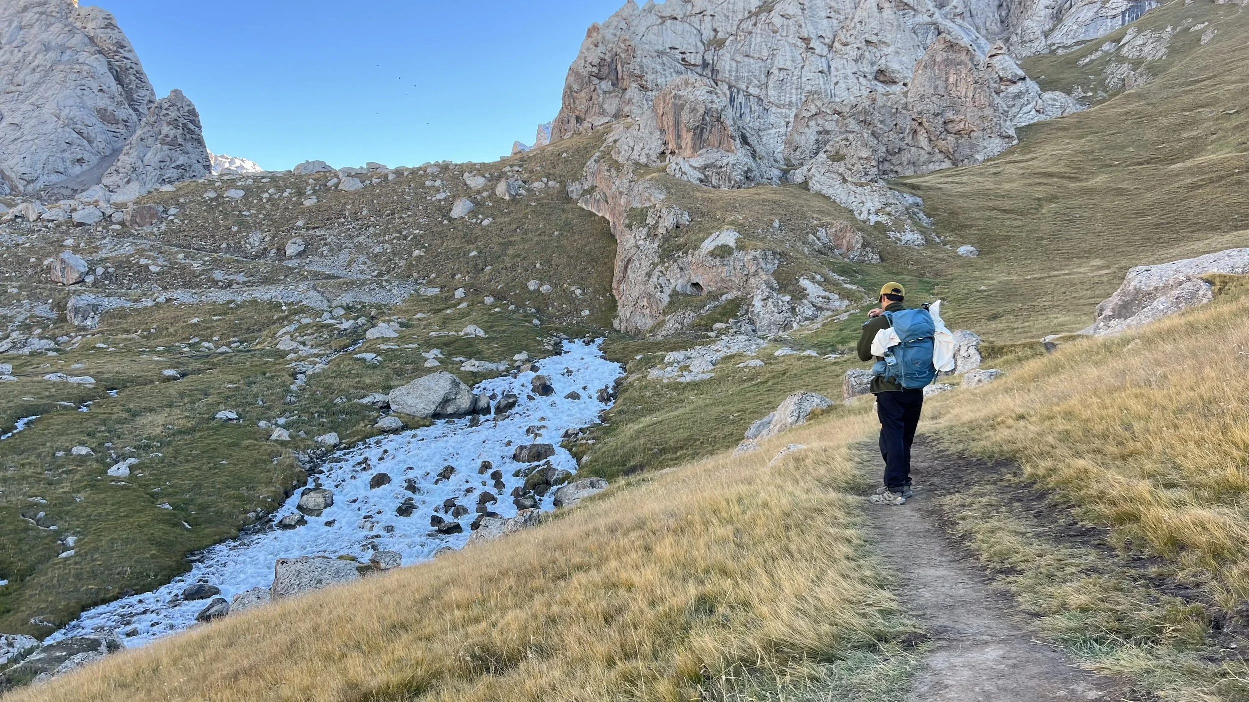 A person with a backpack and cap walking along a mountain trail with rocky cliffs and a small stream in the background.
