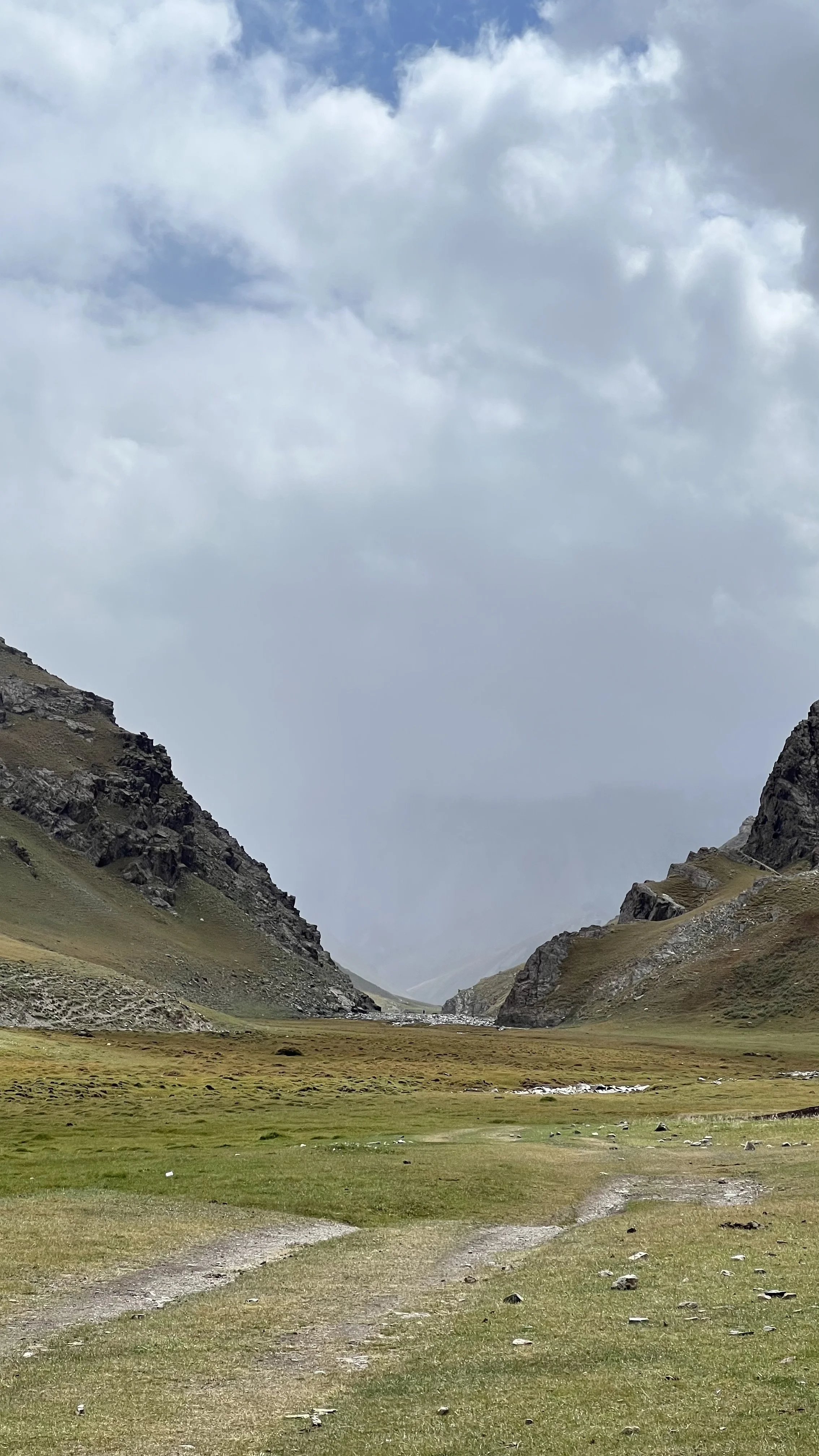A wide valley between mountains with green grass, rocky terrain, and a cloudy sky overhead.