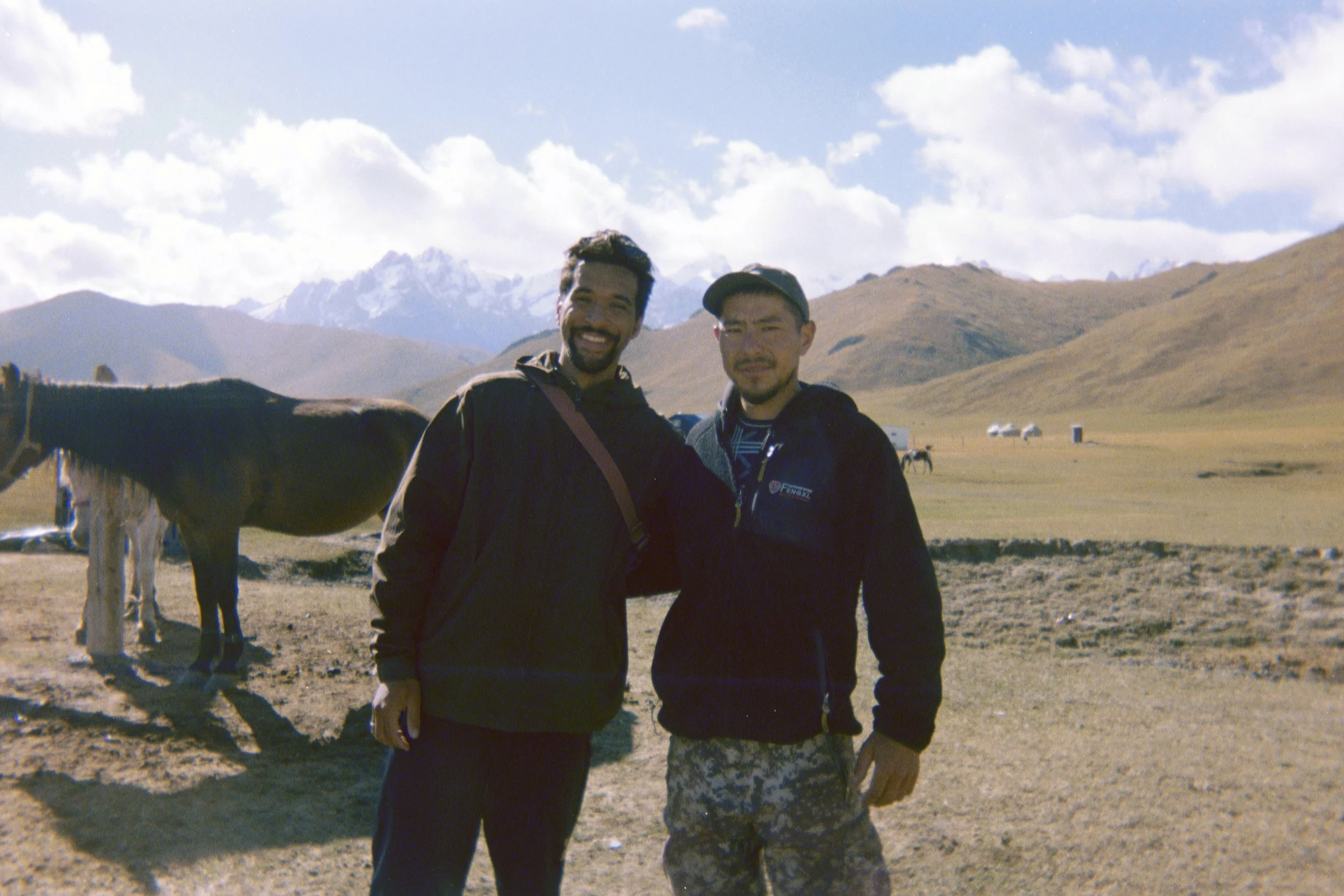 Two men standing outdoors in a mountainous region with a dog partially visible on the left. One man has a beard and is smiling, while the other has facial hair and is squinting. Both are wearing dark jackets. The background features rolling hills, snow-capped mountains, and a partly cloudy sky.