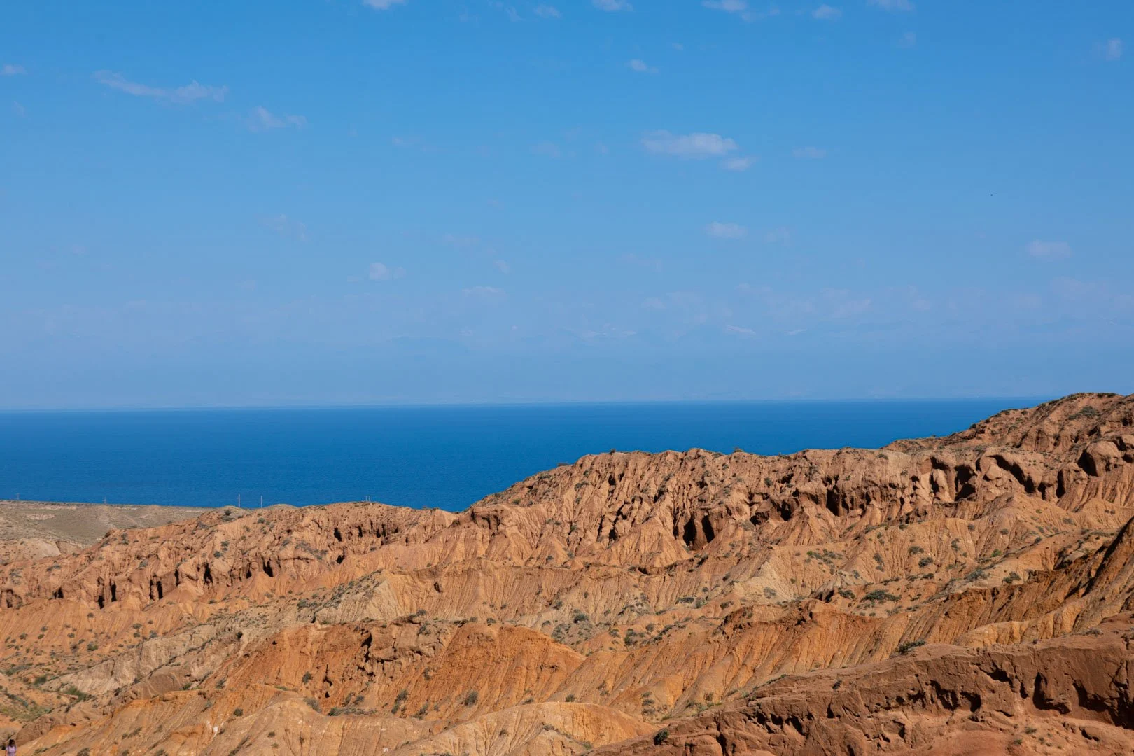 A landscape photo showing orange rocky hills in the foreground and a deep blue ocean in the background, with a clear blue sky.