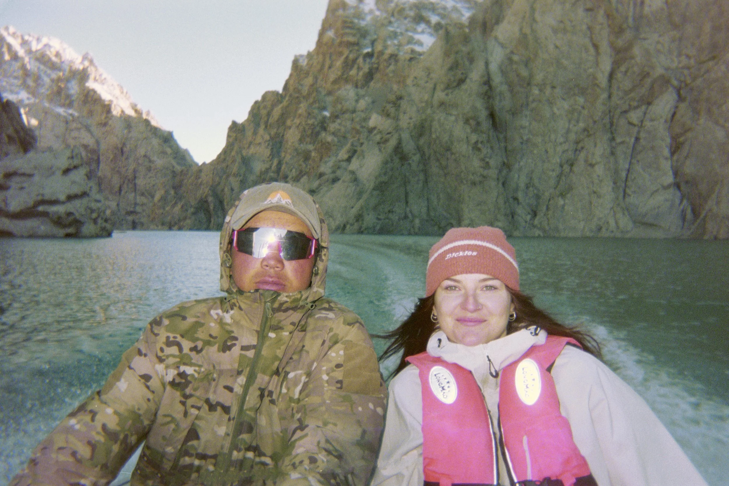 Two people on a boat in a rocky coastal area, one wearing camouflage jacket and sunglasses, and the other wearing a pink life jacket and a red beanie.