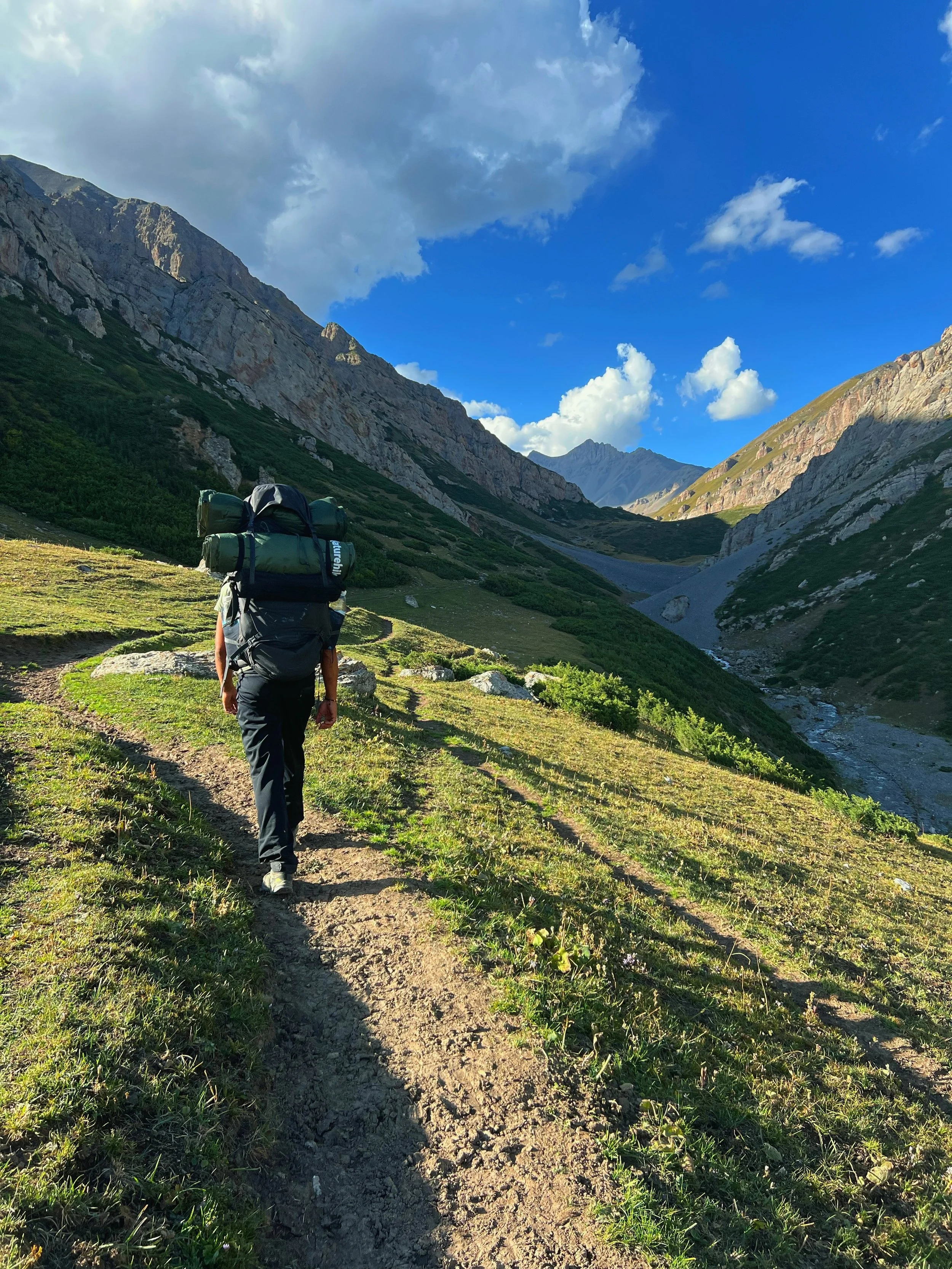 A person with a large backpack hiking on a dirt trail through a mountainous landscape with rocks, green vegetation, and a blue sky with some clouds.