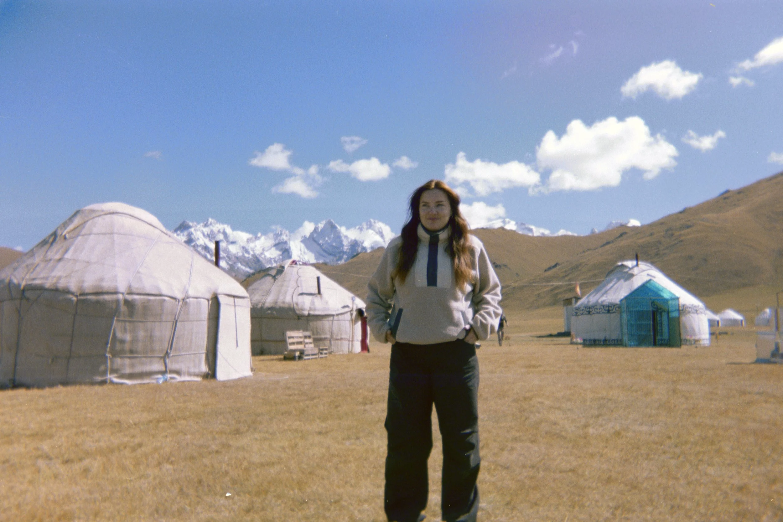 A woman standing in a mountainous landscape with yurts and snow-capped peaks in the background.
