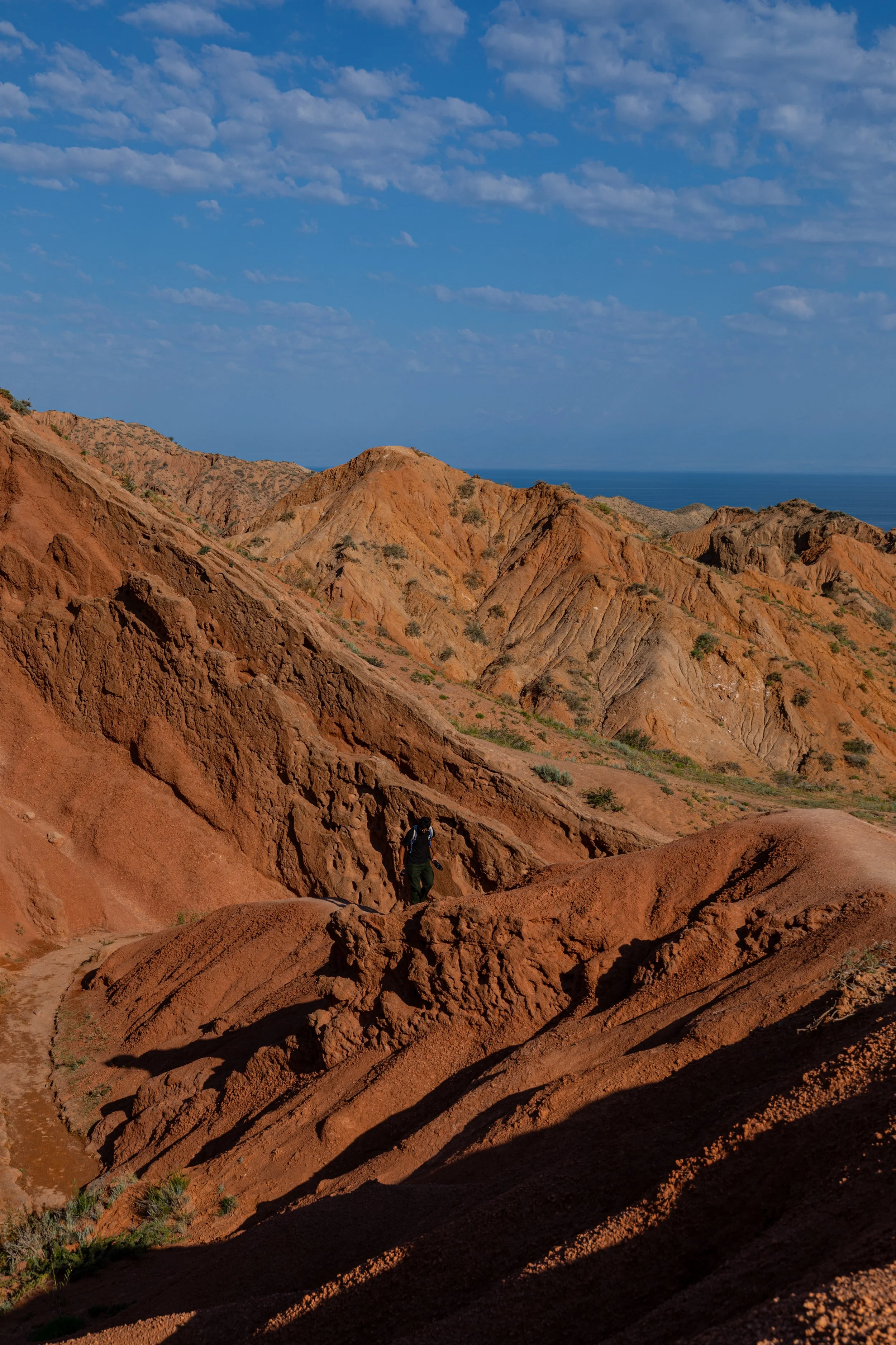 Hiking trail through red clay hills with a person walking and a view of the ocean in the background under a partly cloudy sky.