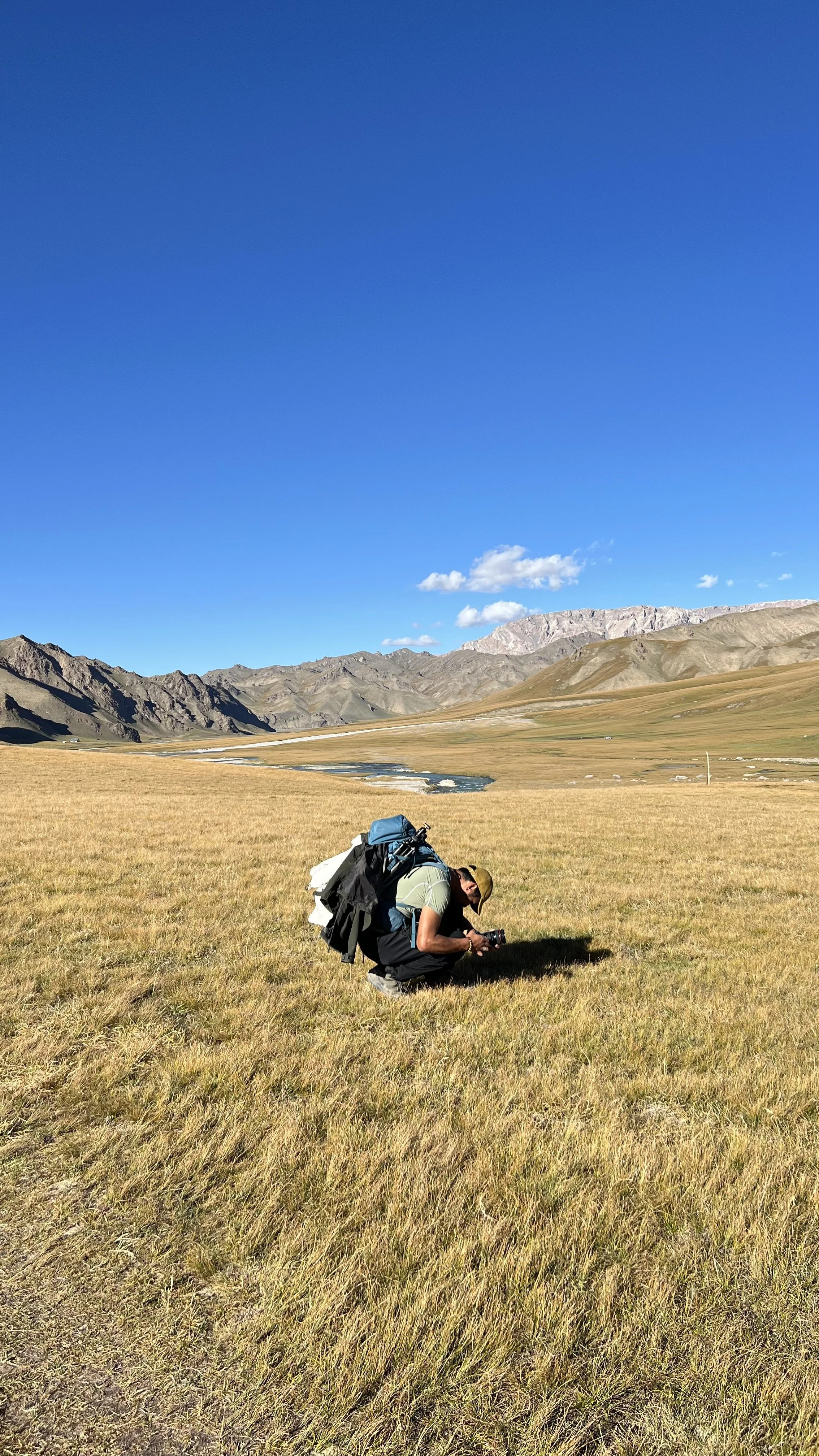 A person kneeling on a grassy plain in a mountainous landscape, taking a photo with a camera. The person has a large backpack and is wearing a cap, with mountains and a river in the background under a bright blue sky.