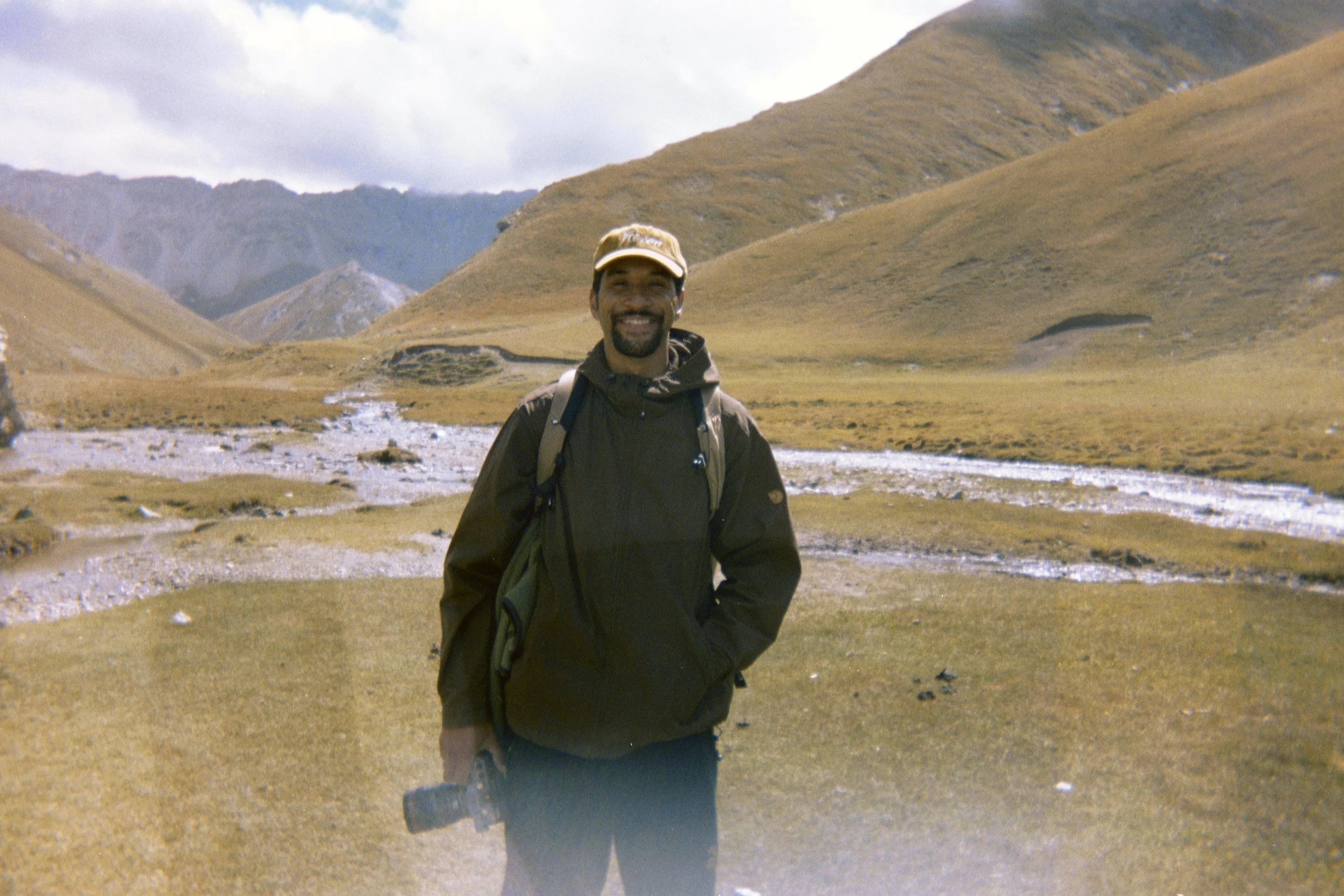 A man smiling while standing outdoors in a mountainous area with a river and grassy hills in the background.