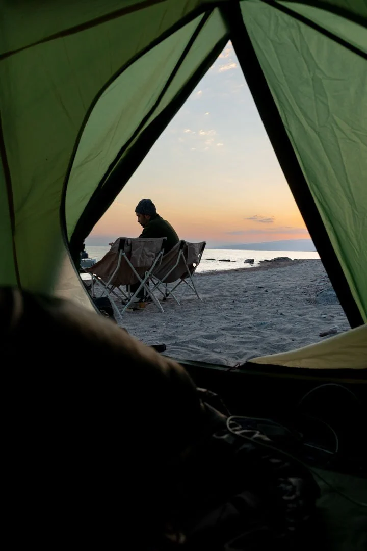 Person sitting in camping chairs on a beach at sunset, viewed from inside a green tent.