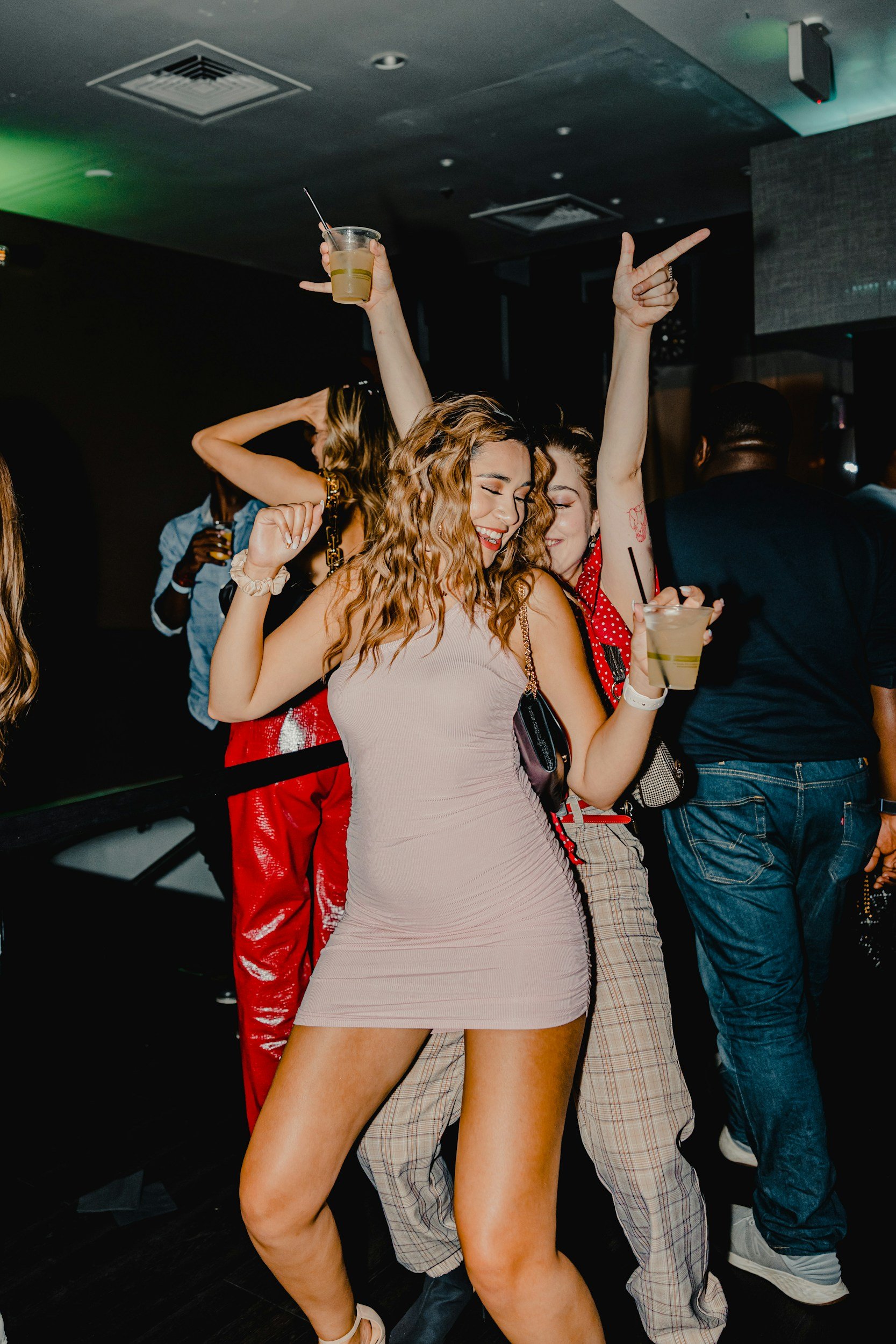 People dancing and enjoying drinks in a nightclub or party setting, with a woman in a beige dress and another woman in a red outfit prominently featured.