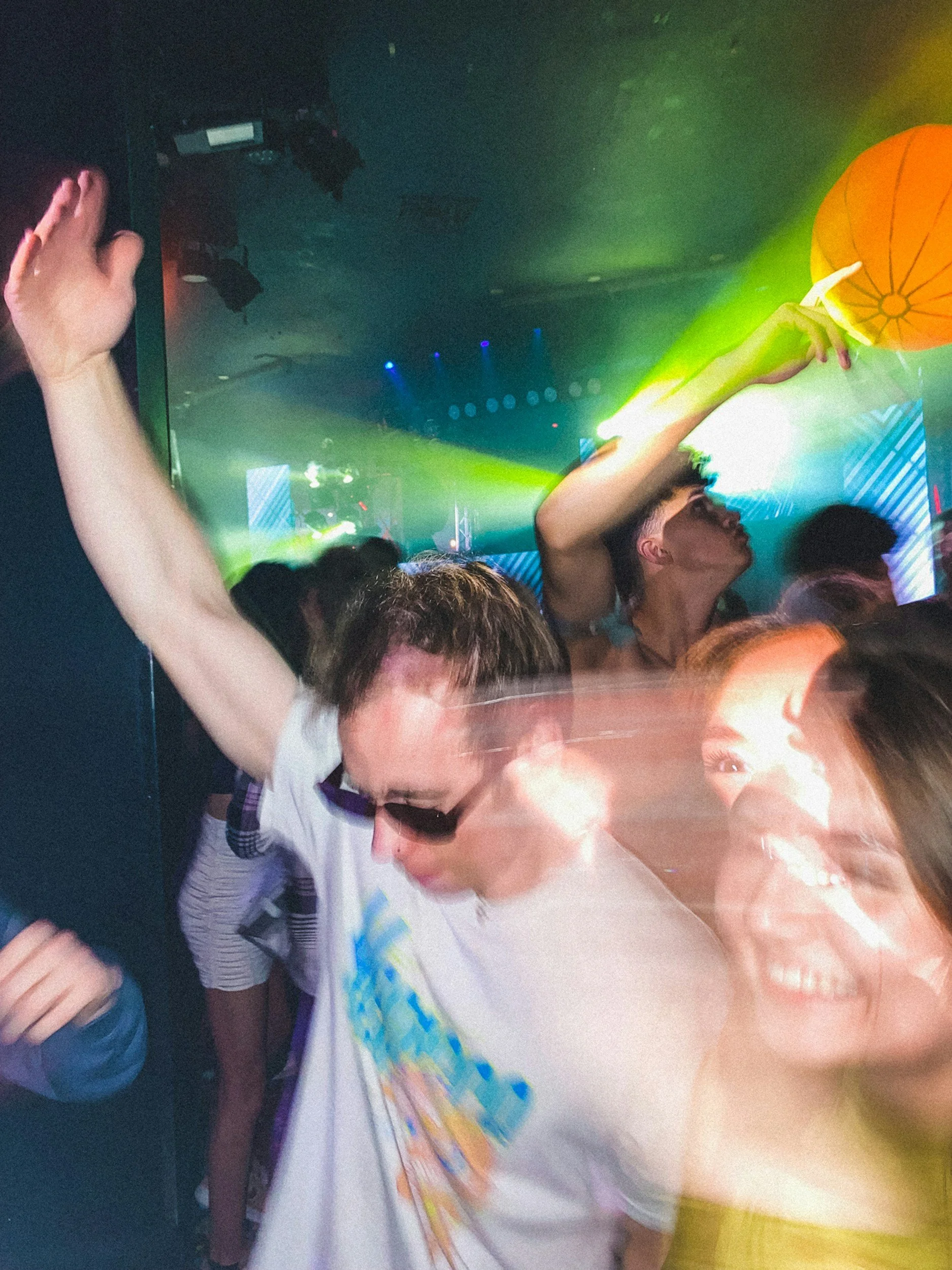 A crowded dance floor with people dancing under colorful lights, including a woman with short hair and a white t-shirt near the center and a woman with decorated face and white clothing in the foreground.