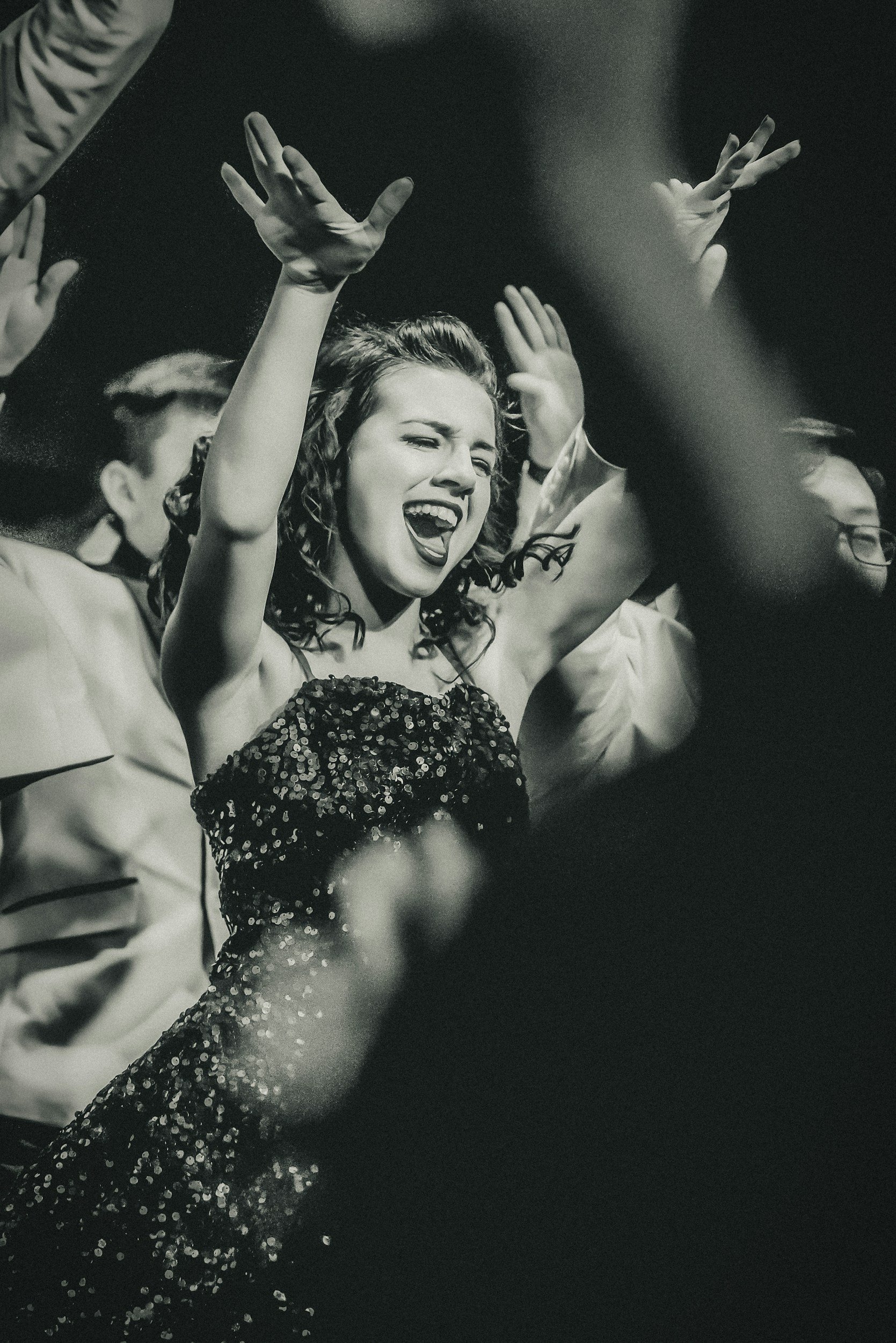 Black and white photograph of a woman with curly hair singing with her eyes closed, wearing a sparkly dress, surrounded by other people with their hands raised in the air.