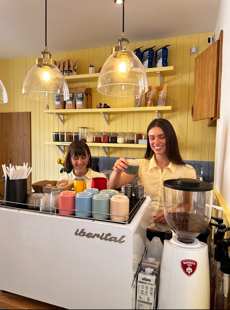 Two women working behind the counter of a coffee shop, with colorful cups, jars of ingredients, and a coffee grinder visible.