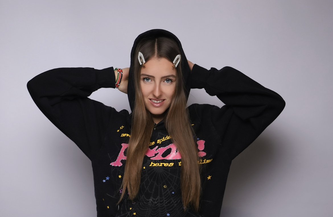 Young woman with long brown hair wearing a black hoodie with colorful text and graphics, adjusting her hoodie against a plain gray background.