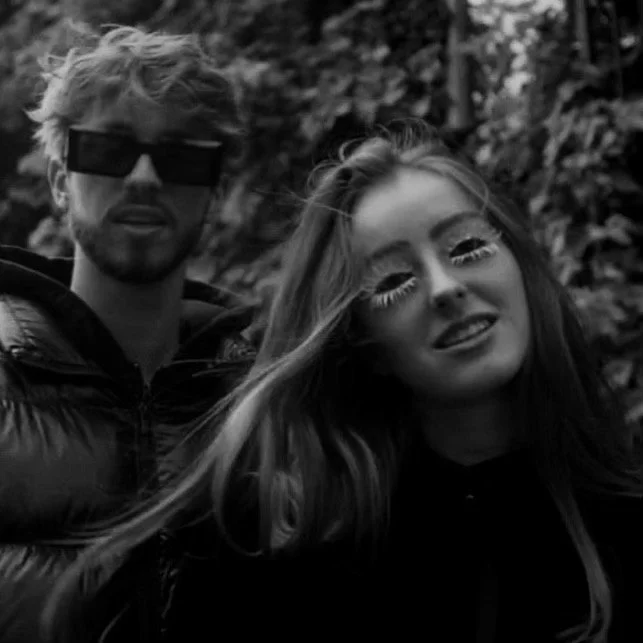 A black and white photo of a young man and woman outdoors, with the man wearing sunglasses and a jacket, and the woman with long hair and makeup.