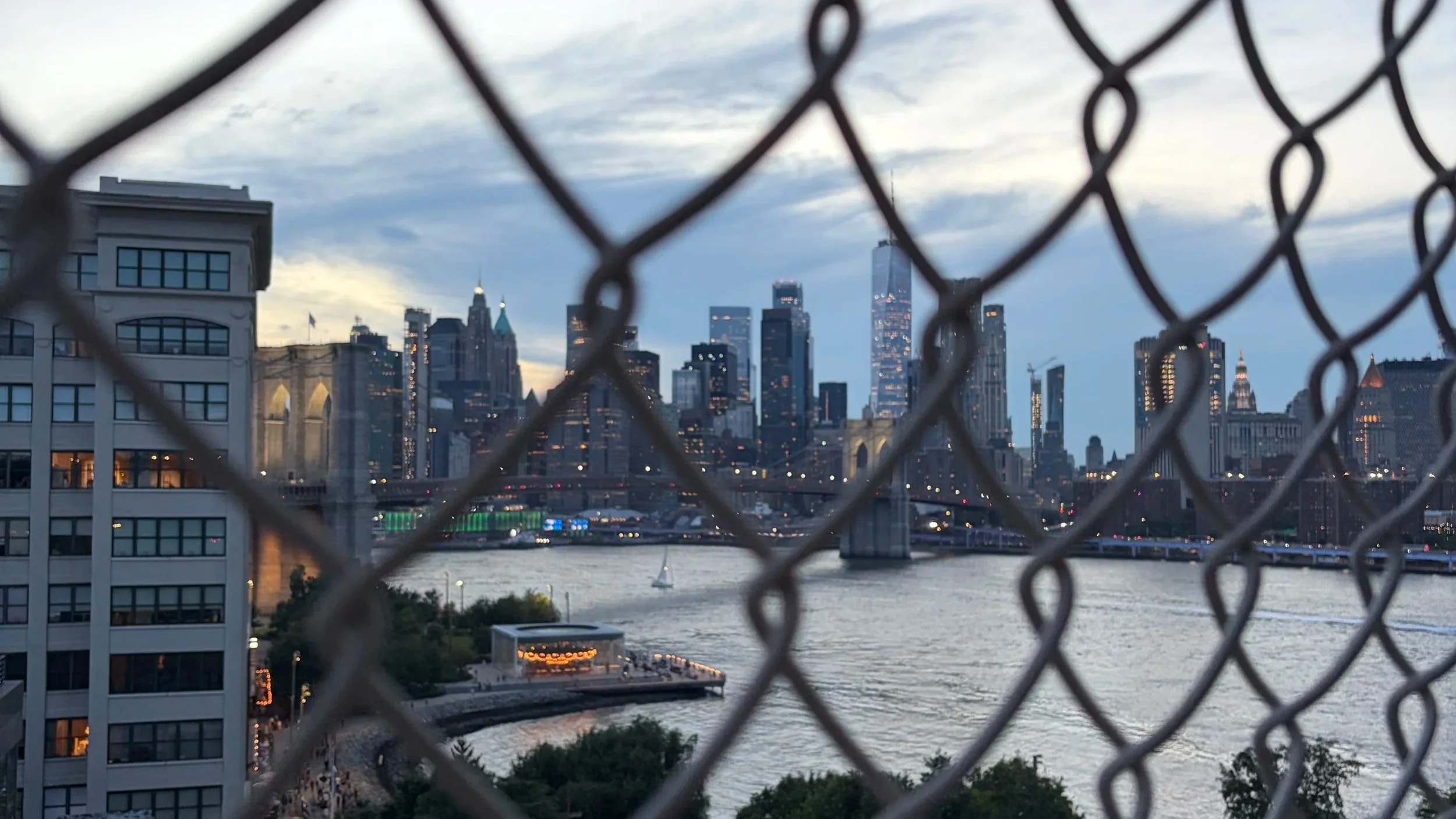 View of the New York City skyline through a chain-link fence, with skyscrapers, river, and bridge in the background.