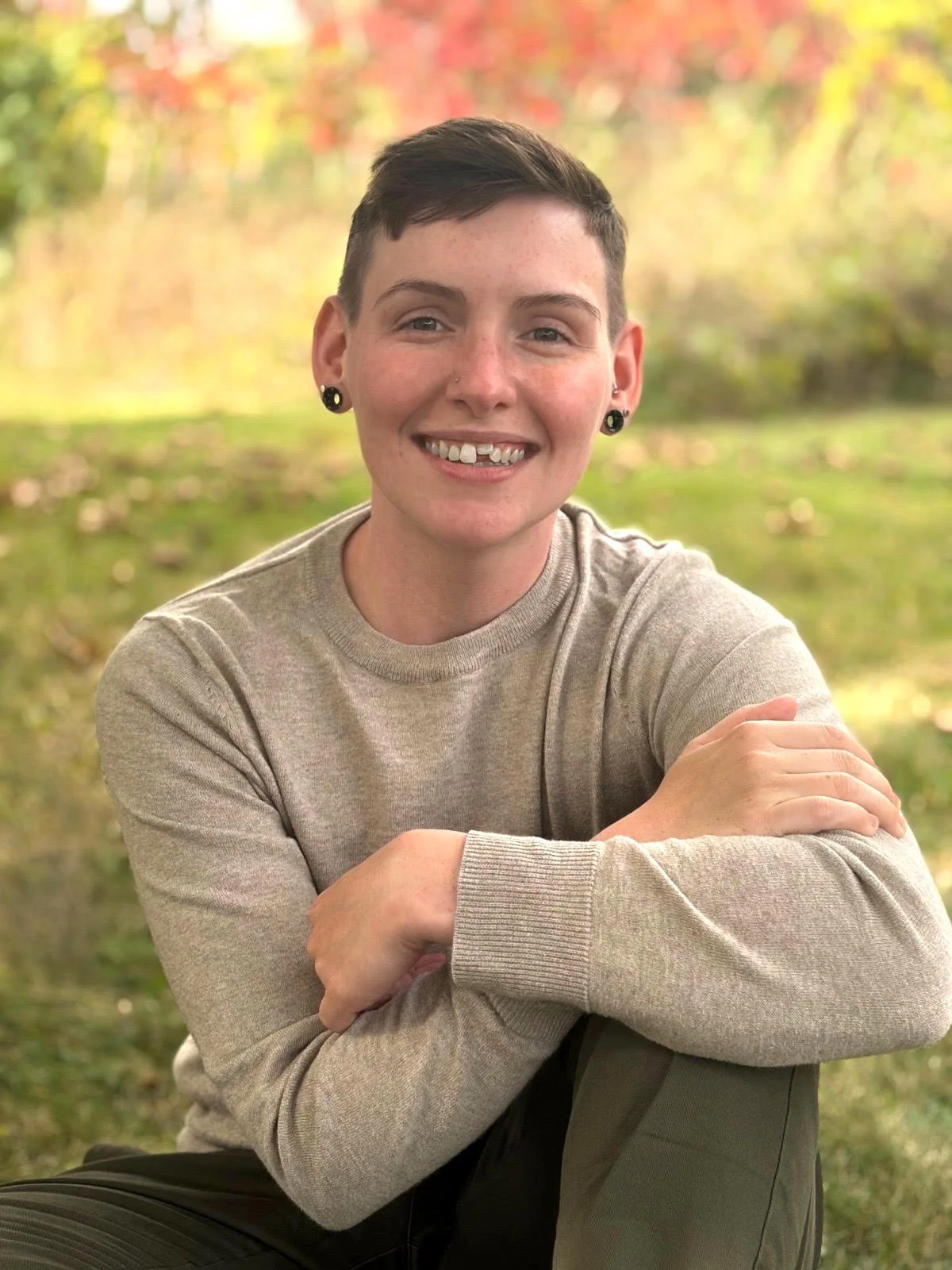 A young person with short dark hair, pierced ears, and a nose ring, smiling and sitting outdoors with arms crossed, in front of blurred autumn foliage.