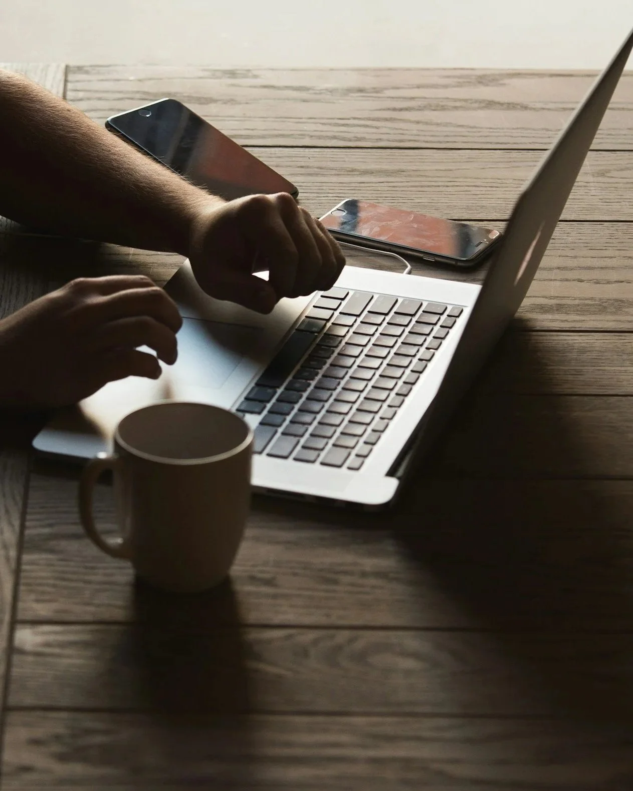A person working on a silver laptop on a wooden table, with two smartphones nearby, a coffee mug in the foreground, and a light-colored wall in the background.