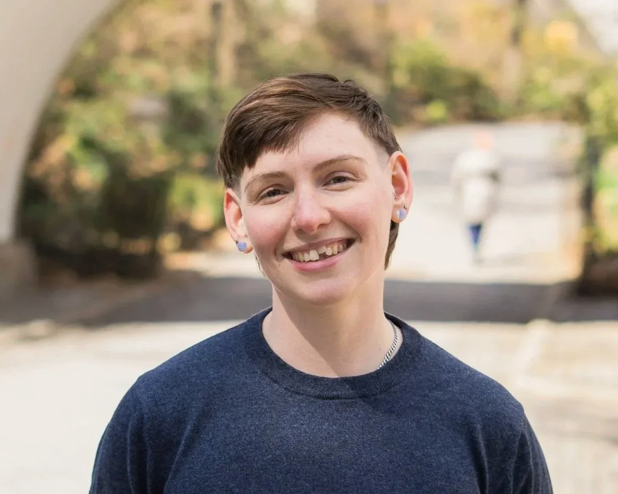 A smiling young woman with short brown hair, wearing a dark blue sweater and small earrings, standing outdoors with blurred trees in the background.