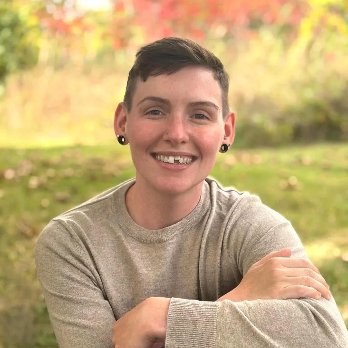 A smiling person with short dark hair and earrings, sitting outdoors with a background of trees with fall foliage.