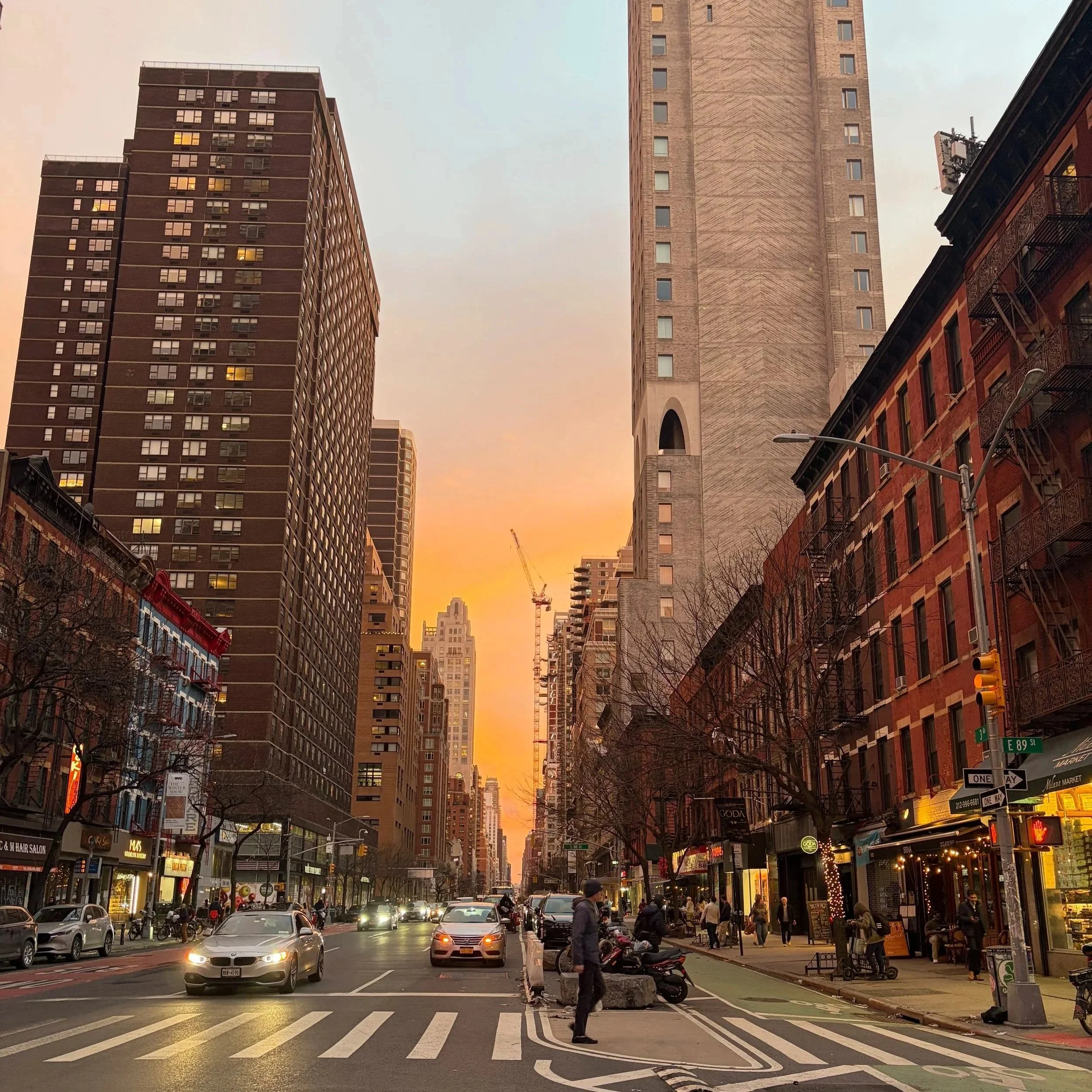 City street scene during sunset with tall buildings, shops, cars, pedestrians, and streetlights.