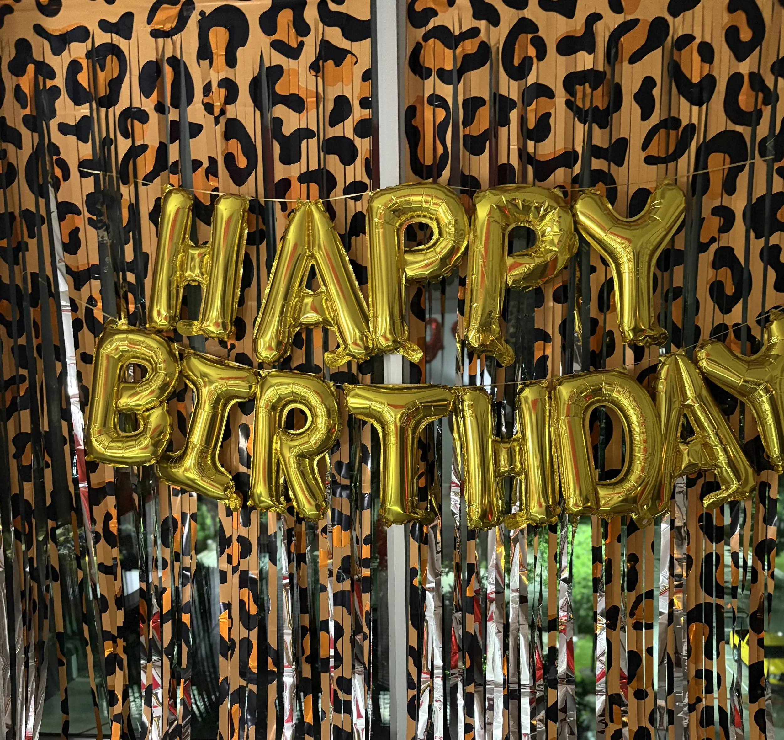 Gold foil balloons spelling “HAPPY BIRTHDAY” against an orange and black leopard-print party backdrop.