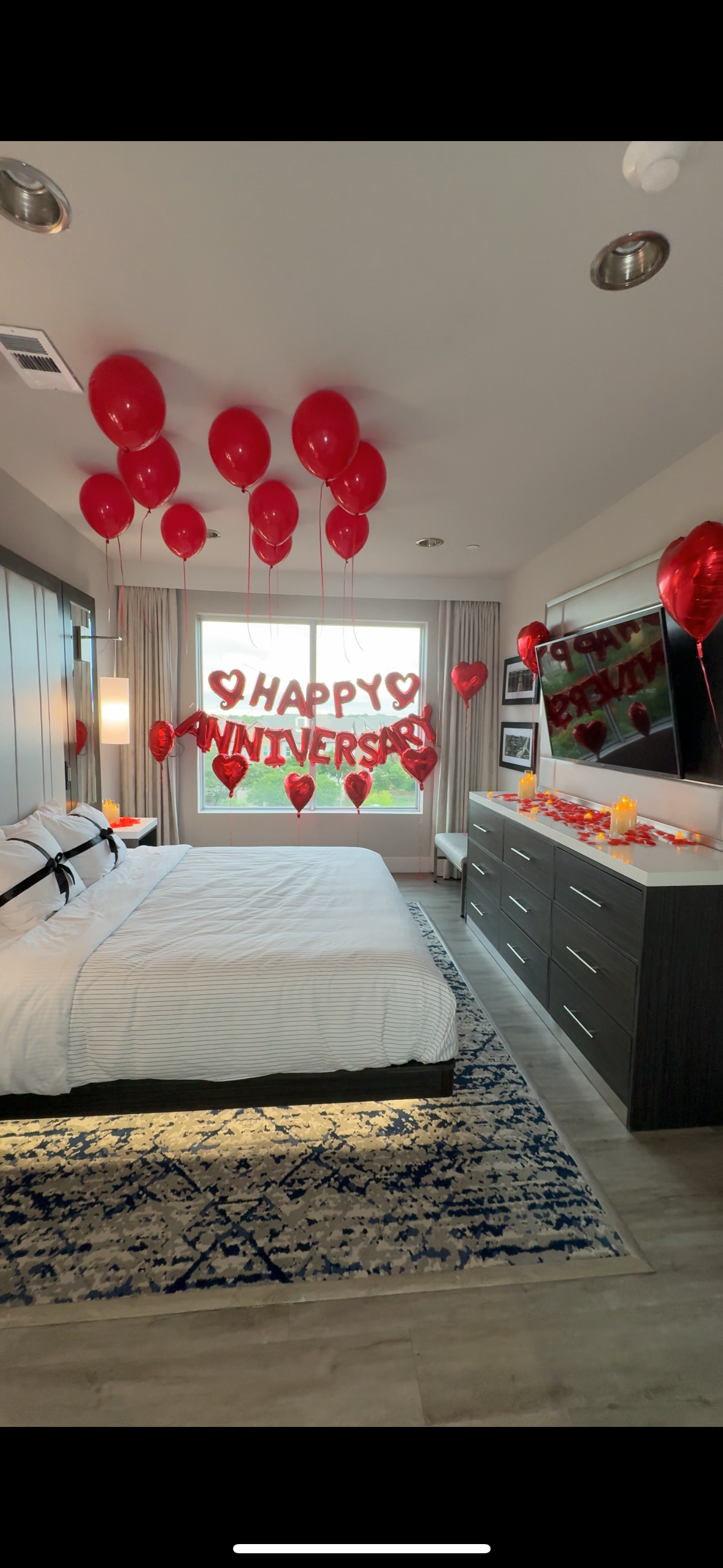 Romantic bedroom anniversary setup with red balloons, heart-shaped balloons, candles, rose petals, and a “Happy Anniversary” sign.