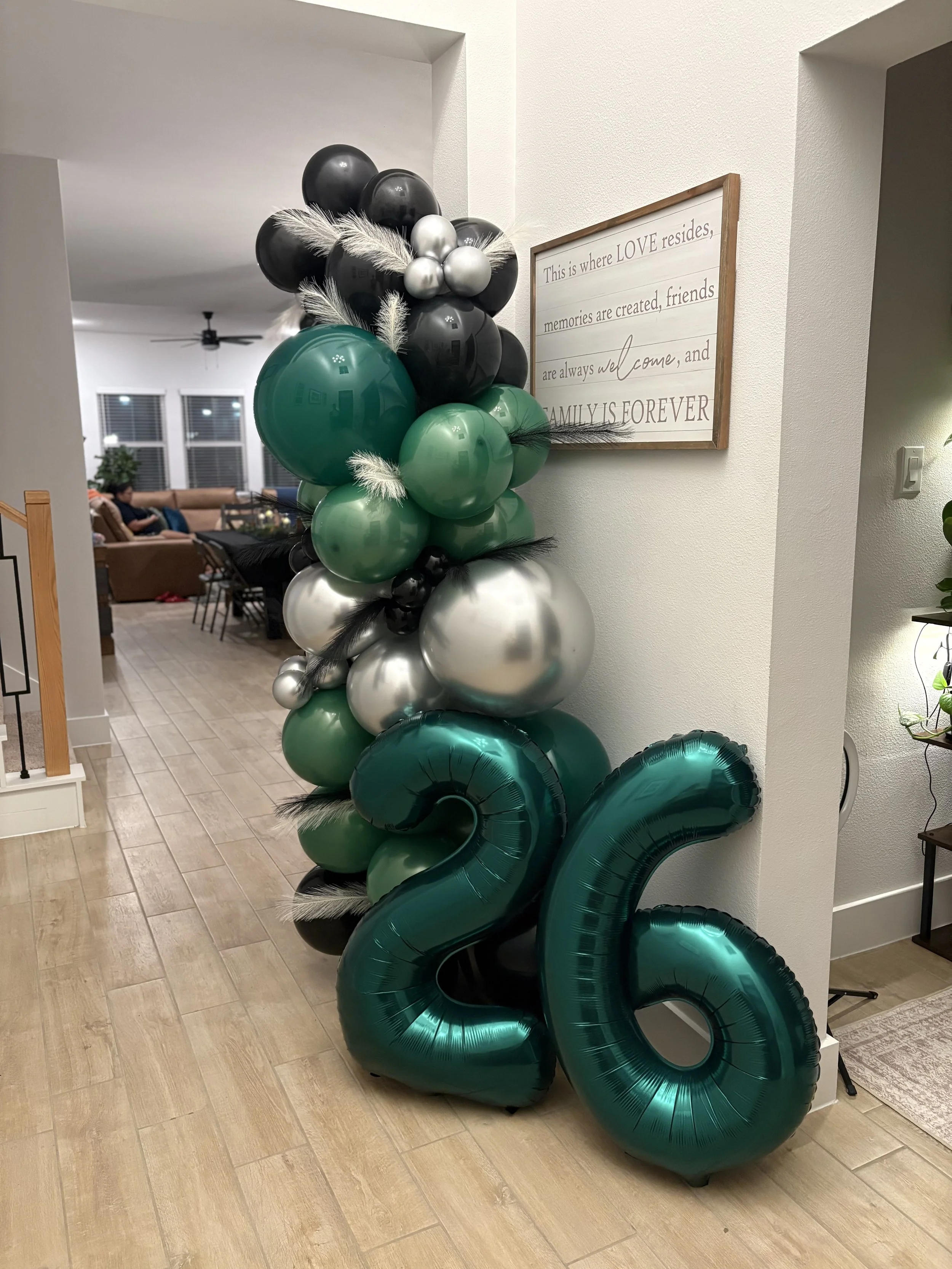 Large black, green, and silver balloon arrangement with decorative feathers beside a framed family quote sign in a home